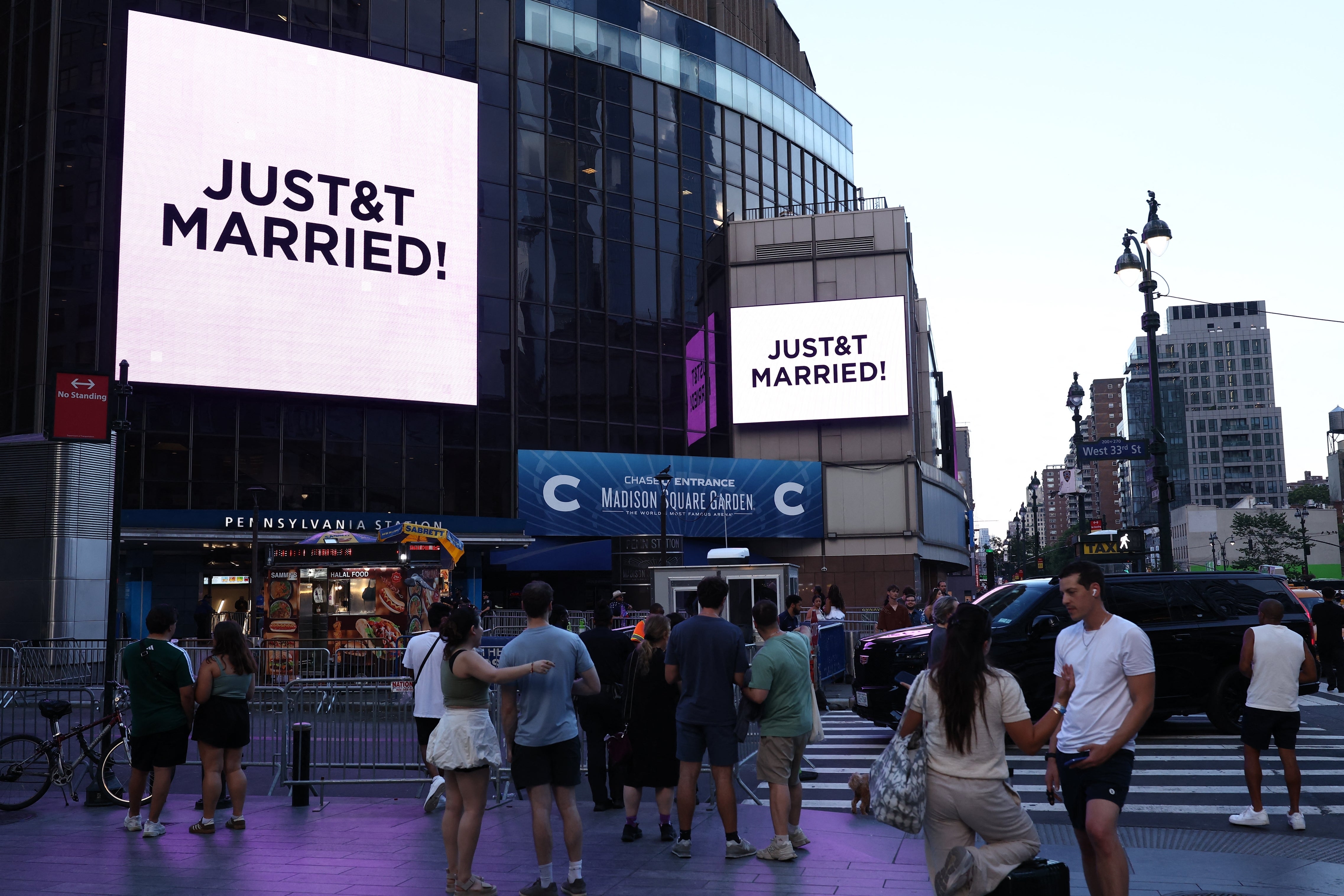 Large digital screens in a busy city area display the message "JUST&T MARRIED!" as people walk by