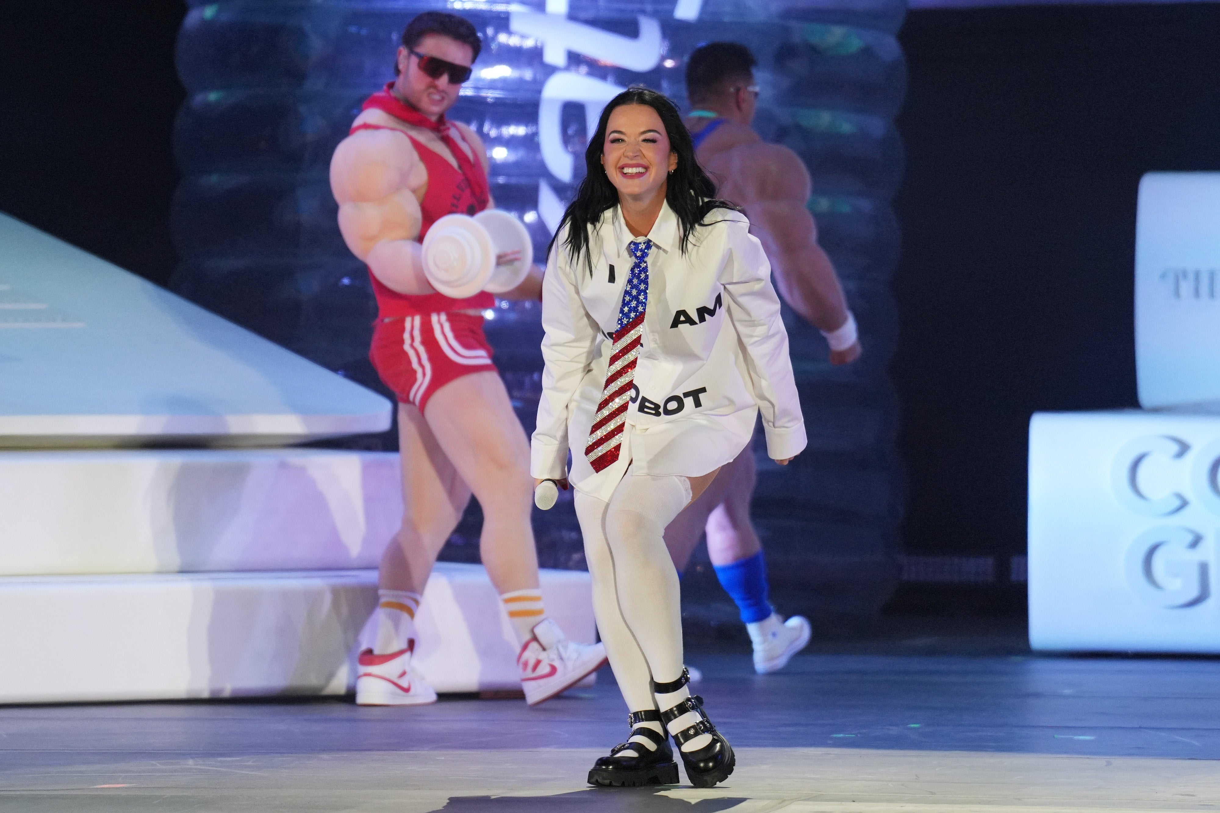 Katy Perry in a white outfit with "I am a robot" text, performing on stage with inflatable weights and others in athletic wear as background elements