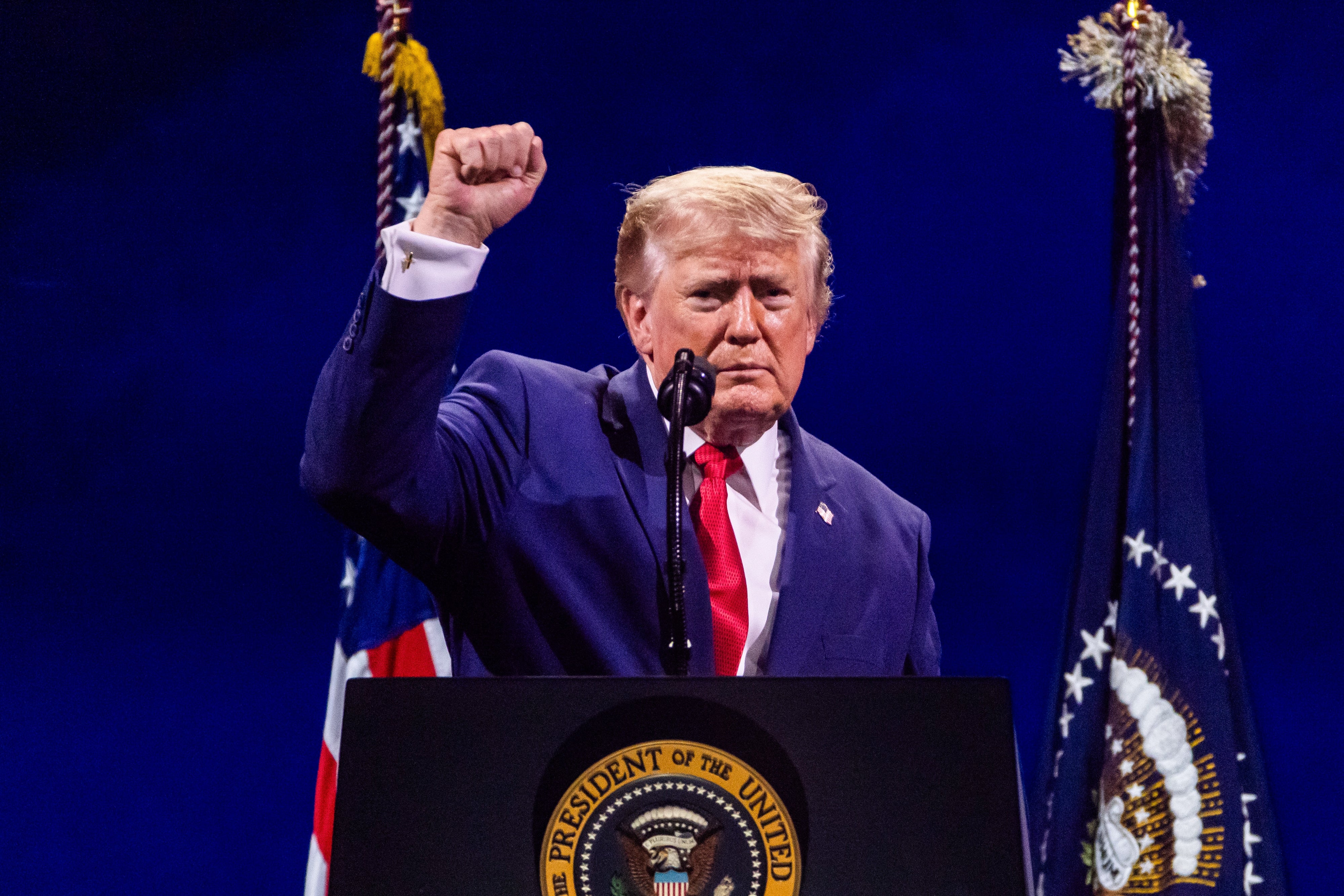 A antheral successful a suit stands astatine a podium with a statesmanlike seal, raising his fist. Two flags are disposable successful the background