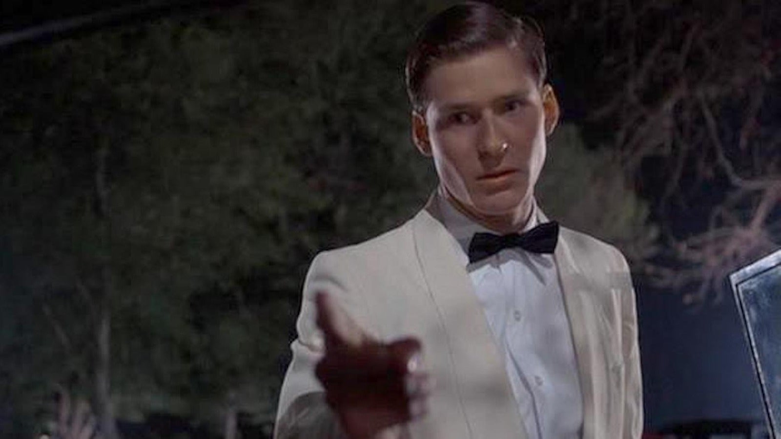A person in a formal white suit with a bow tie gestures outward against a dimly lit outdoor backdrop