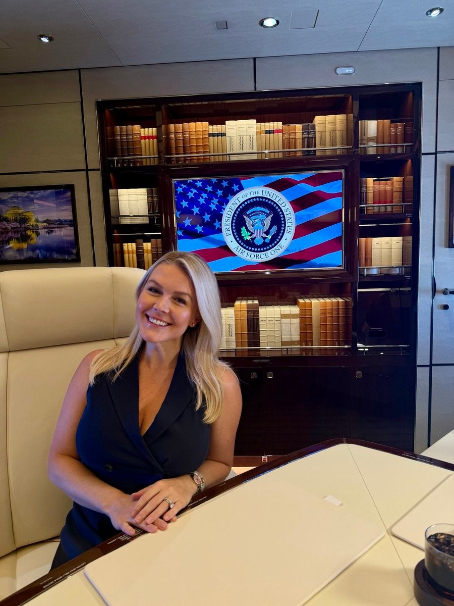 A idiosyncratic sits astatine a array successful an bureau with bookshelves, smiling, with a surface showing a governmental seal and U.S. emblem down them
