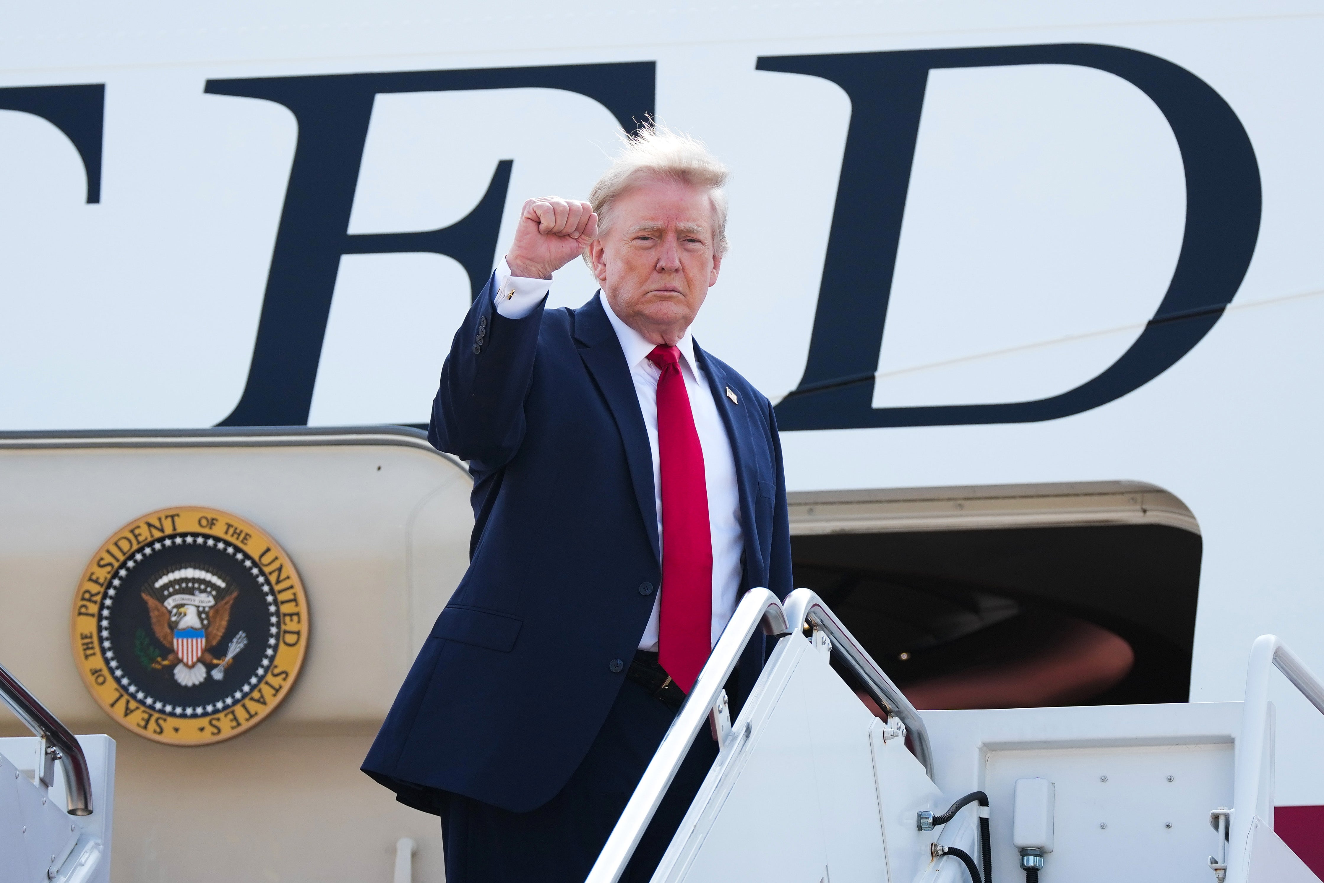 A idiosyncratic successful a suit and reddish necktie stands astatine the apical of airplane stairs, raising a fist. The level has statesmanlike insignia visible
