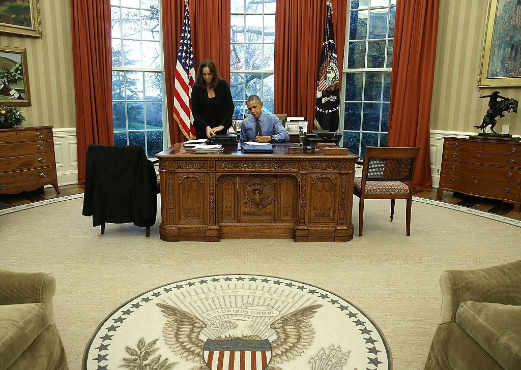 Person som sitter ved Resolute Desk med en annen stående i nærheten, begge i formell antrekk, inne i det ovale kontoret med flagg og presidentsegl synlig
