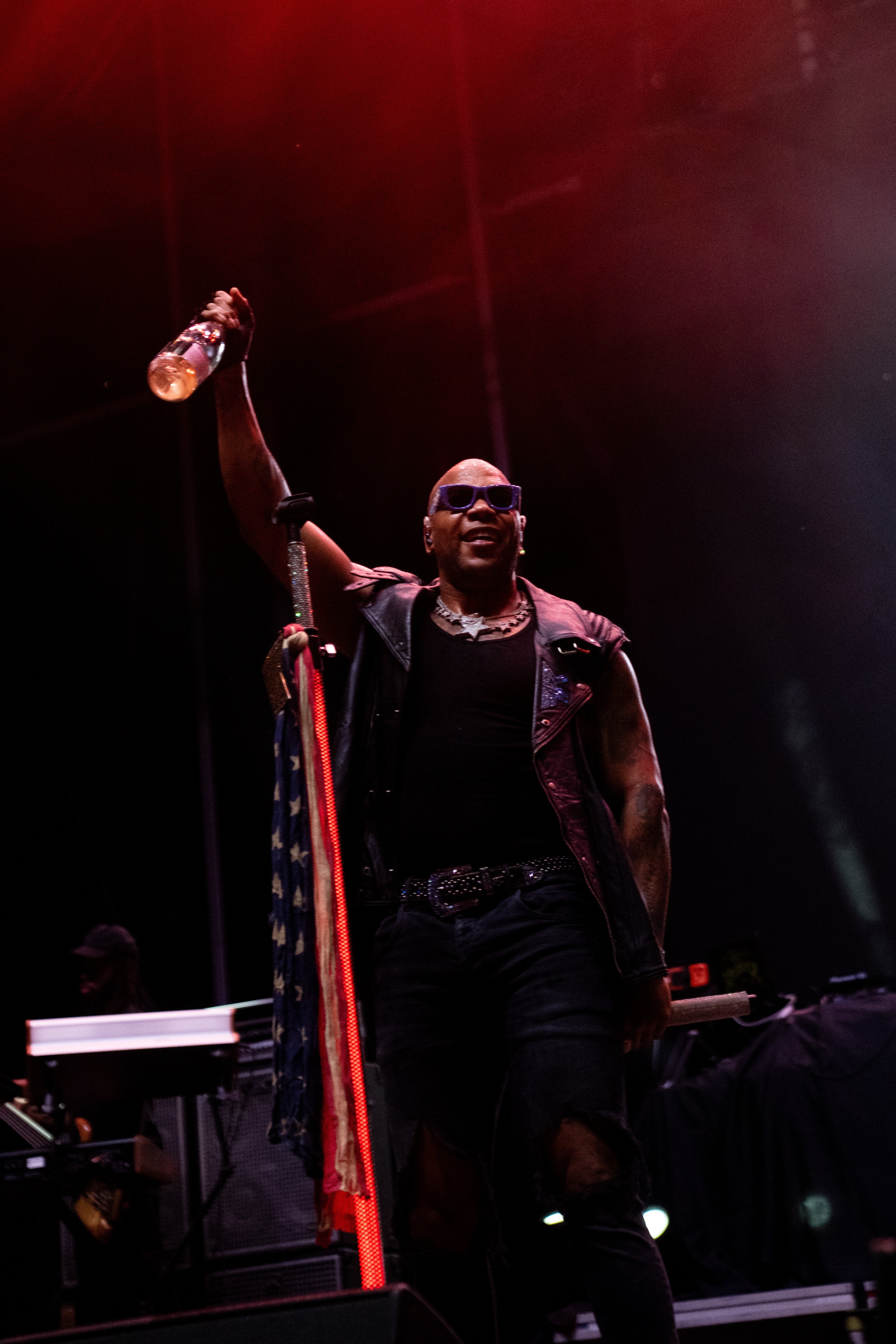Person connected signifier holds vessel aloft, wearing a leather vest and sunglasses, performing energetically astatine an outdoor concert