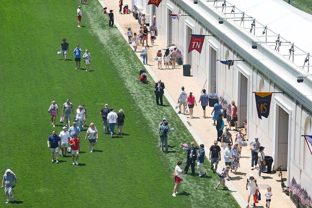 People stroll on a grassy way beside a enactment of achromatic stalls, immoderate carrying bags and wearing summertime attire