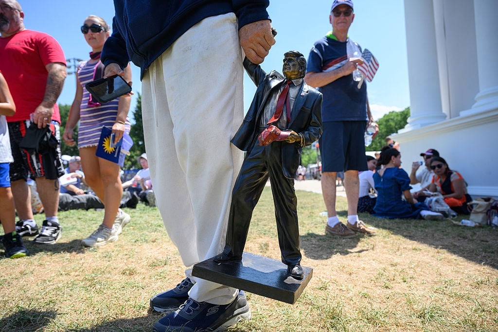 A idiosyncratic holds a tiny statue of a antheral successful a suit portion others basal and beryllium adjacent connected a grassy country during a nationalist event