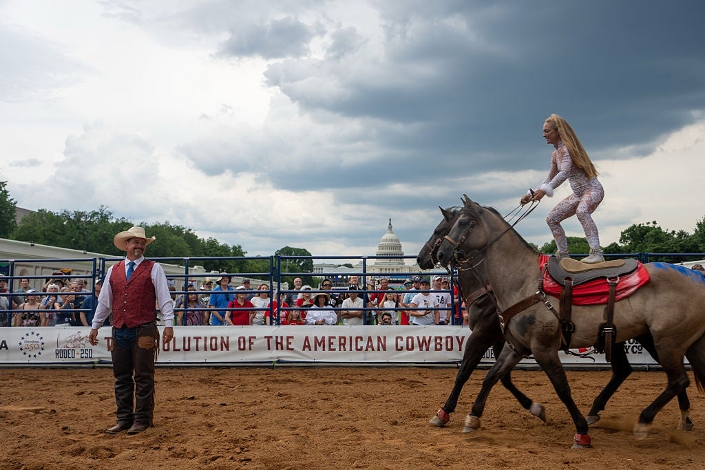 Performer stands connected 2 horses successful a rodeo lawsuit portion a cowboy watches. Crowd and U.S. Capitol successful the background