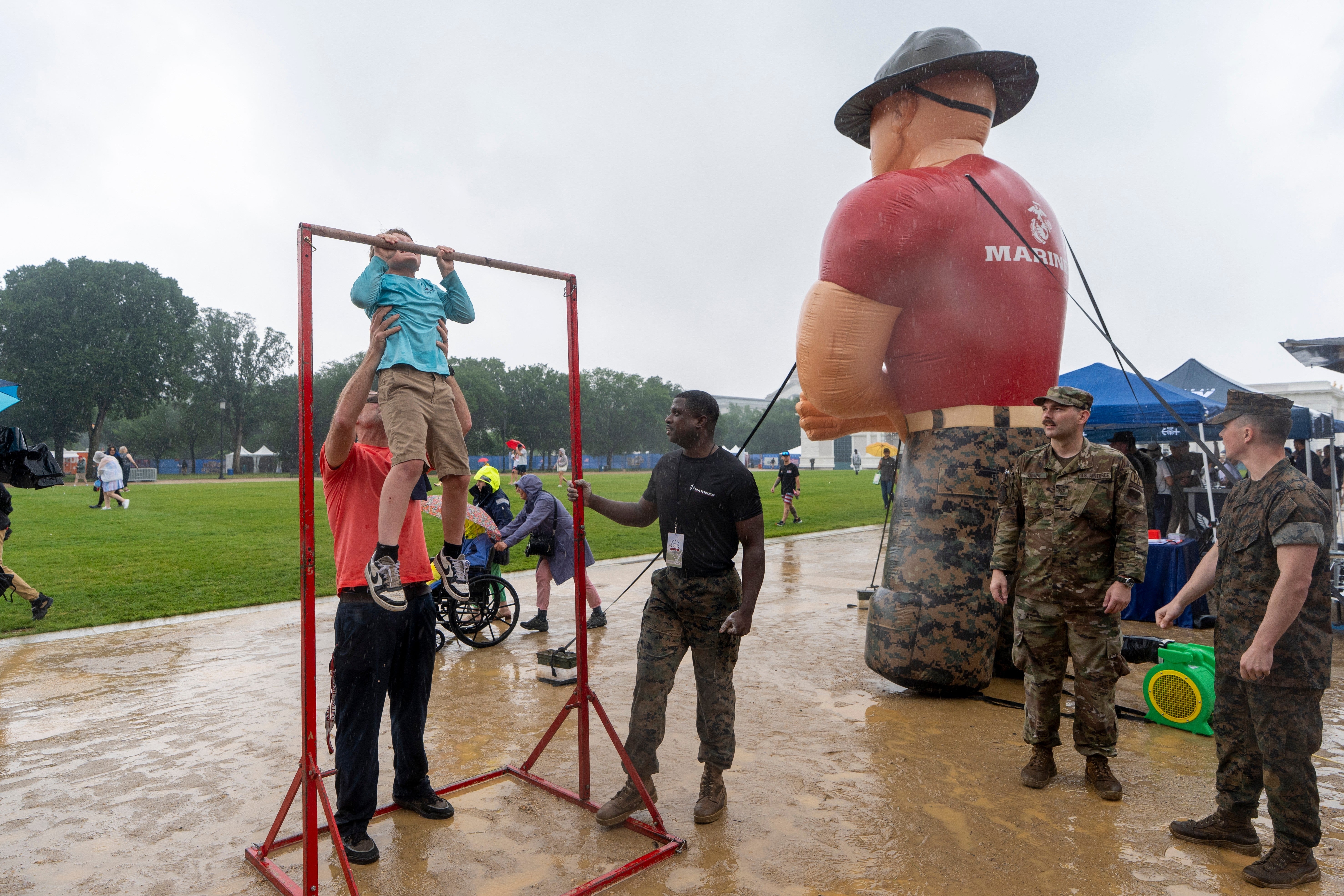 A kid performs a pull-up assisted by a man, portion others, including subject personnel, ticker astatine an outdoor lawsuit with a ample inflatable Marine figure