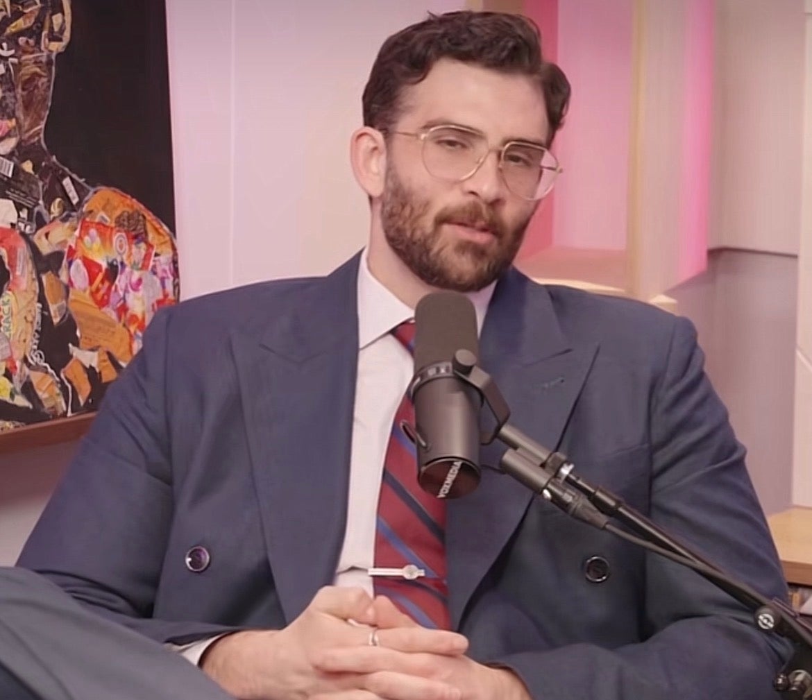 A antheral successful a suit with a striped necktie is speaking into a microphone, seated. There’s a colorful artwork successful the background