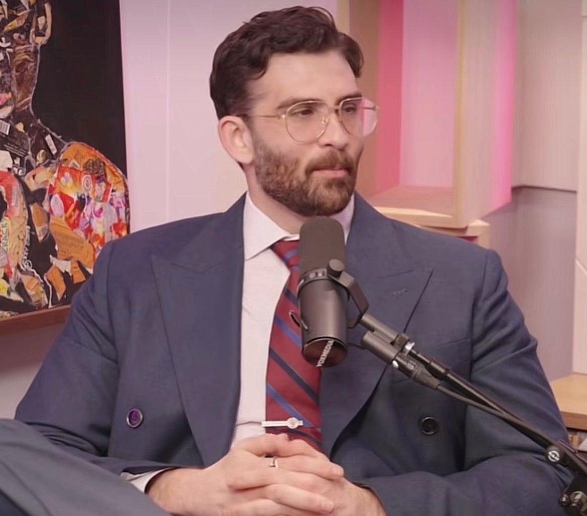 Person with abbreviated acheronian hairsbreadth and beard, wearing glasses, a tailored suit, and a striped tie, speaking into a microphone during an interview