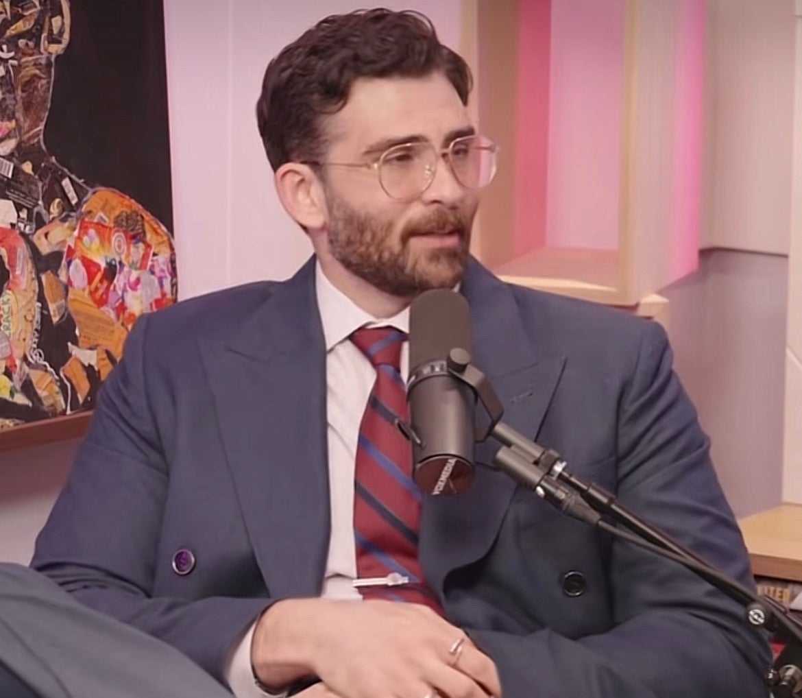 Person successful a podcast studio, wearing glasses, a suit with a tie, and sitting successful beforehand of a microphone
