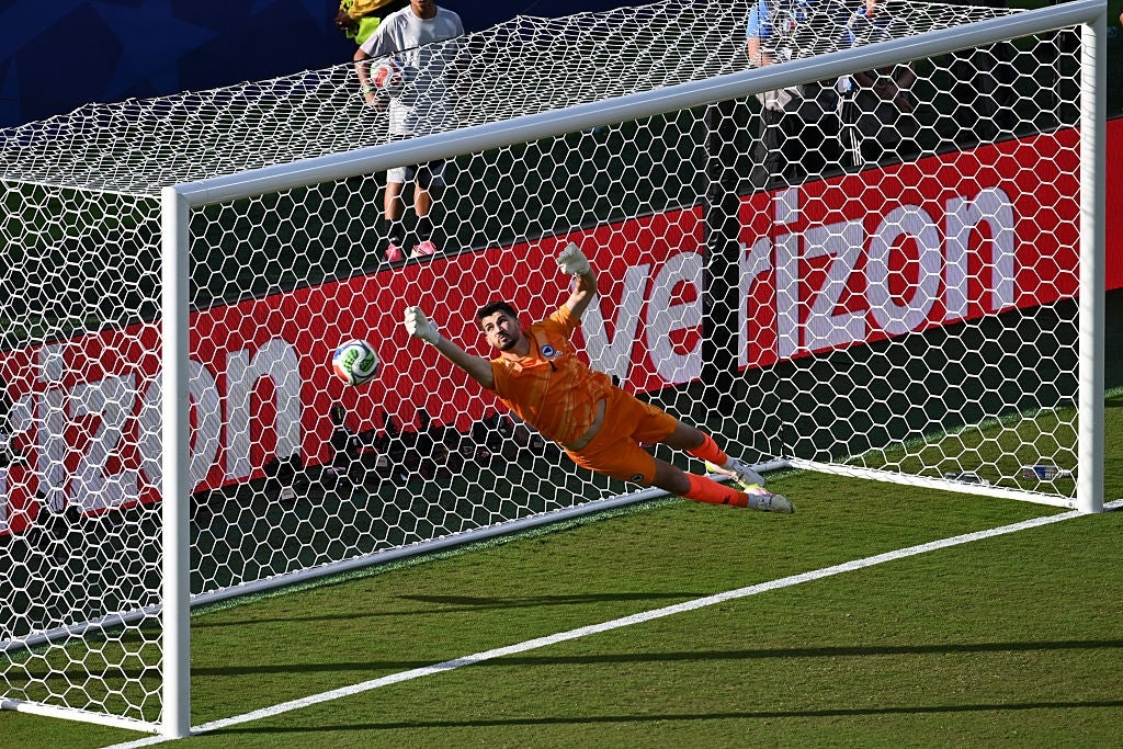 En fotballkeeper i oransje utstyr hopper til venstre og forsøker å blokkere en ball fra å gå inn i målet under en kamp