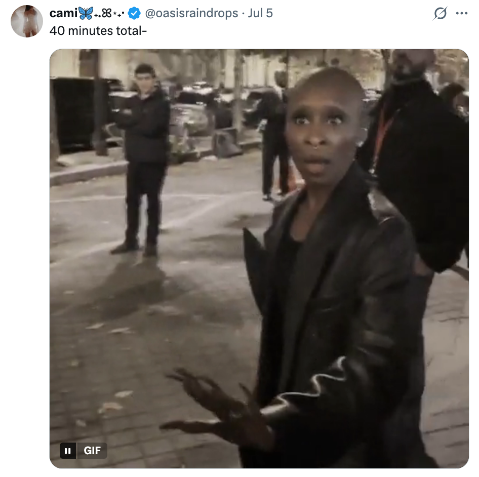A person outdoors gesturing, wearing a stylish leather jacket, with others in the background