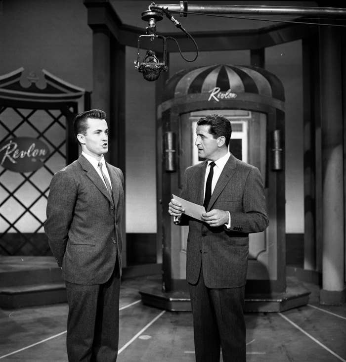Two men in suits on a TV set, one holding paper, likely in conversation or interviewing. Vintage studio style with "Revlon" sign in background