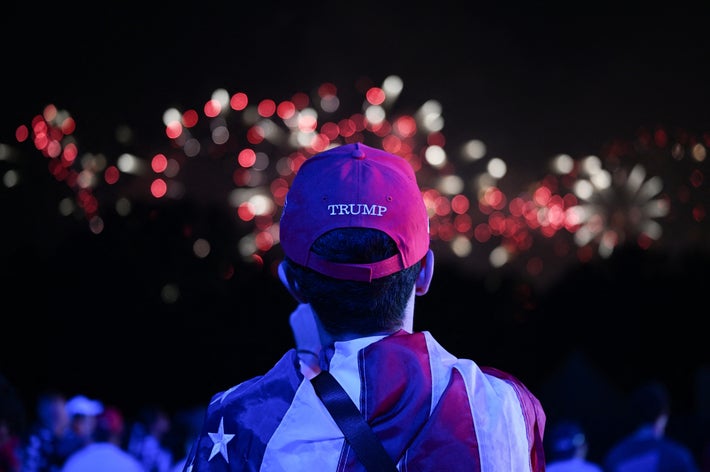 男性が「TRUMP」と書かれた赤い帽子をかぶり、アメリカ国旗柄の服を着て花火を見ている。