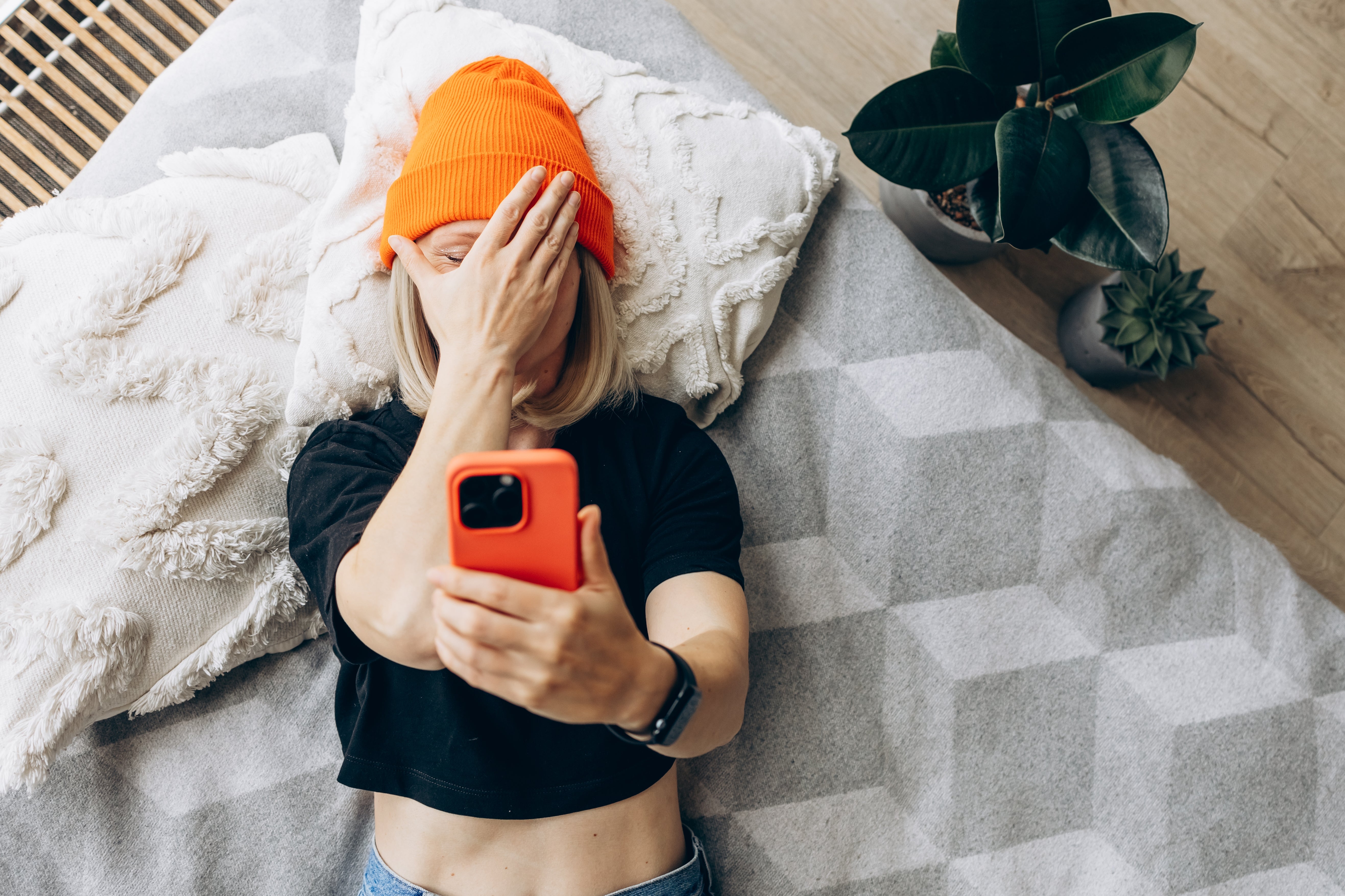 Person lying connected  a furniture  covers their look   with 1  manus  portion    taking a selfie with a smartphone. They deterioration  a beanie and casual clothing