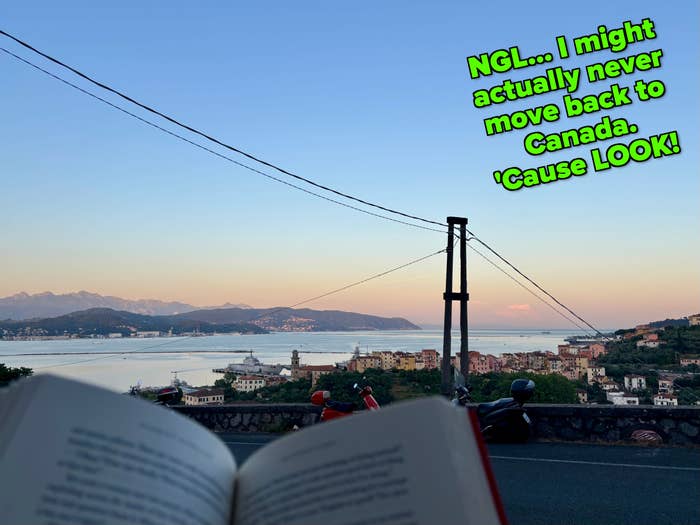 An open book with blurred text is held up against a scenic view of a coastal town with mountains in the background at sunset