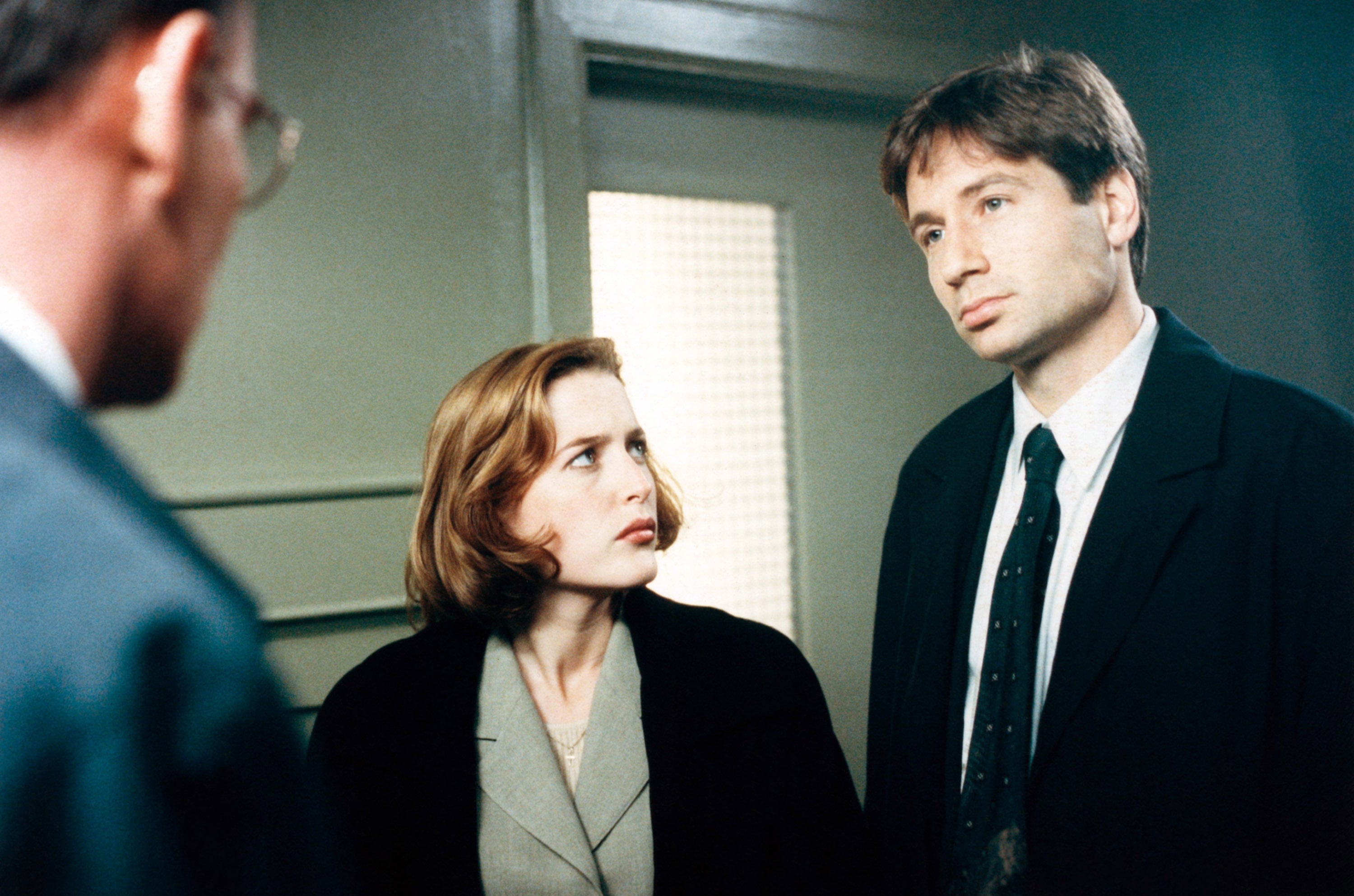 Two detectives in suits are in a serious discussion with a man in a suit. They appear to be inside an office or building hallway