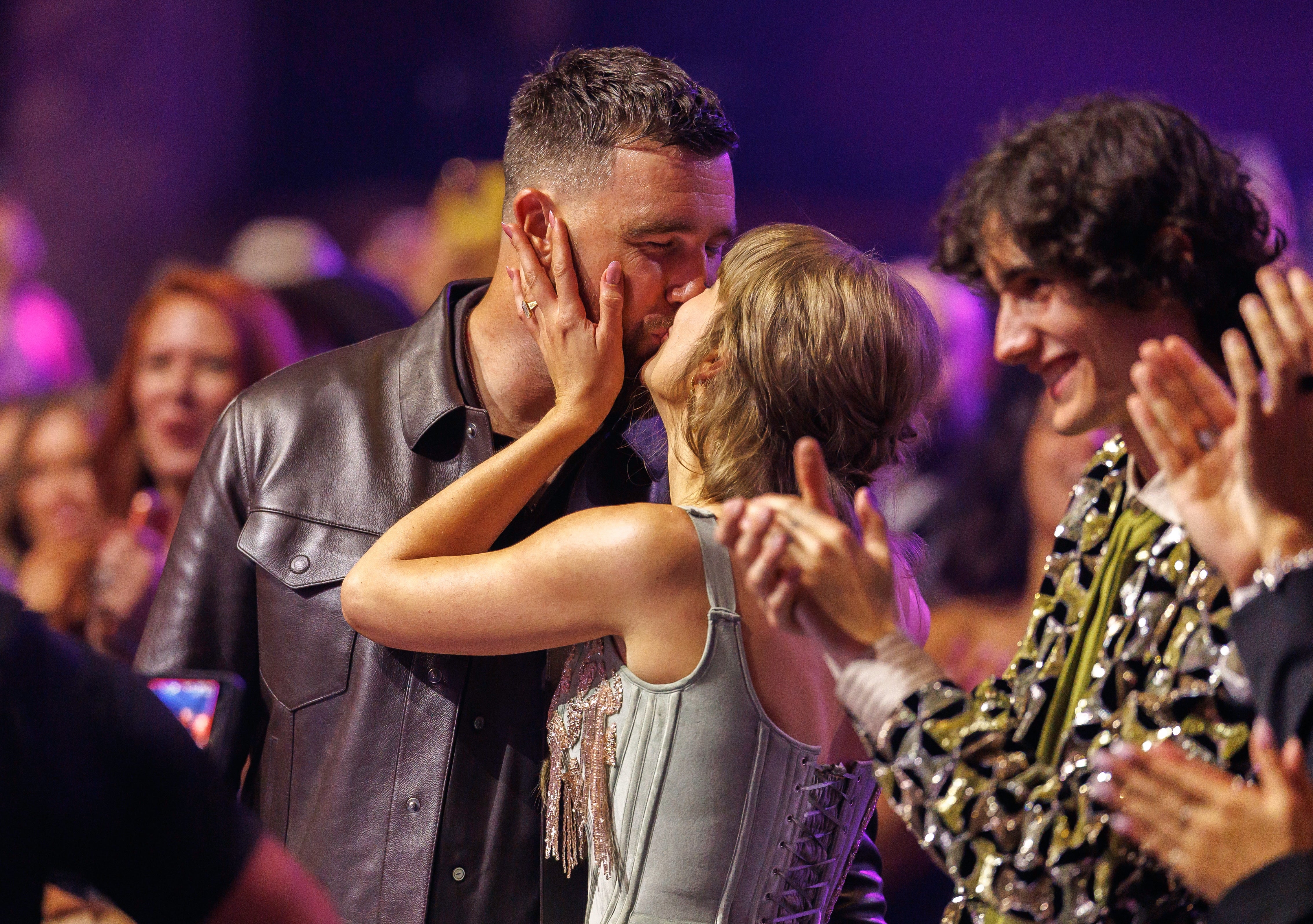A couple shares a kiss at a public event, surrounded by clapping people. The woman wears a fitted dress, and the man is in a leather jacket
