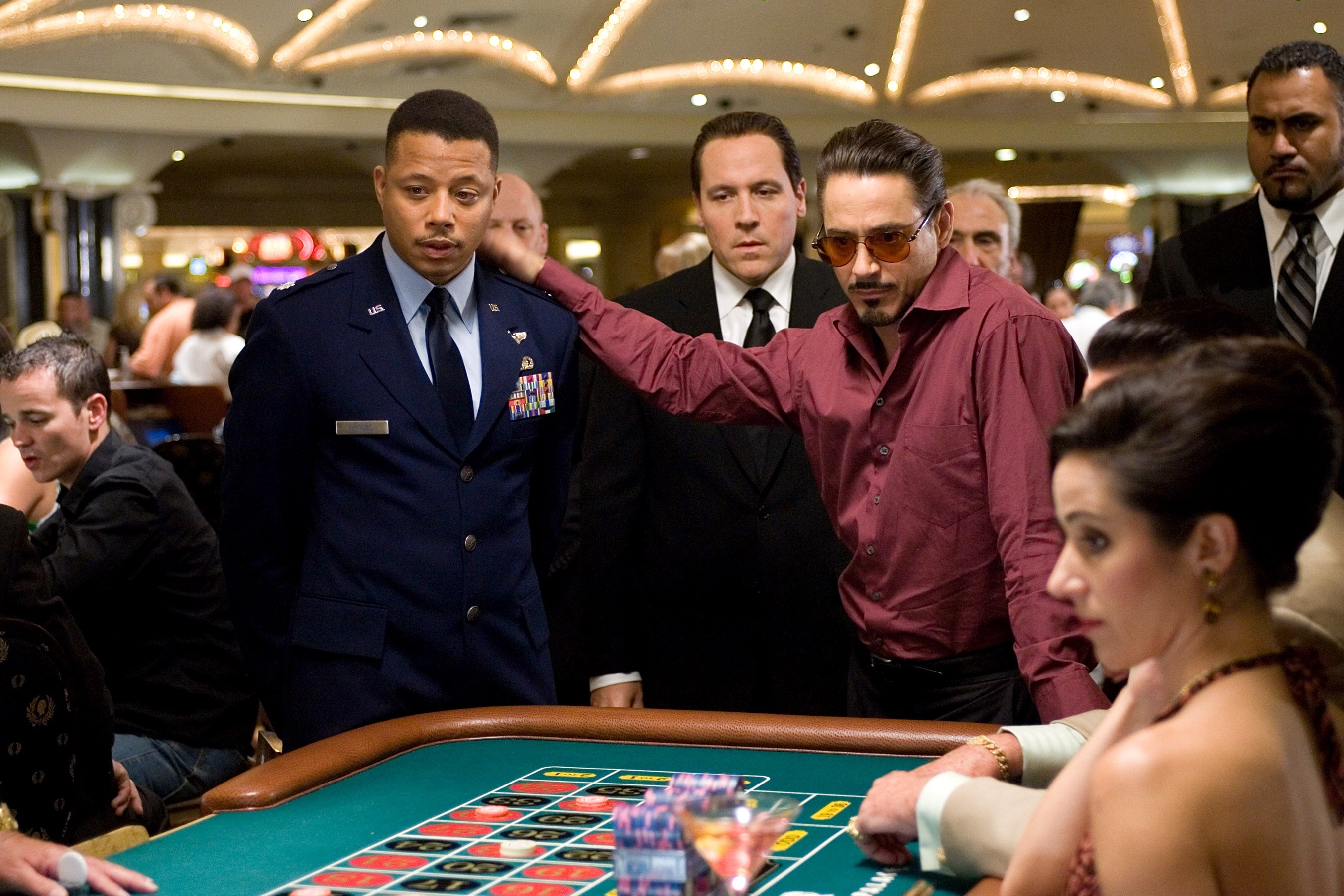 A man in a military uniform and a stylishly dressed man in sunglasses stand at a crowded casino table, surrounded by onlookers