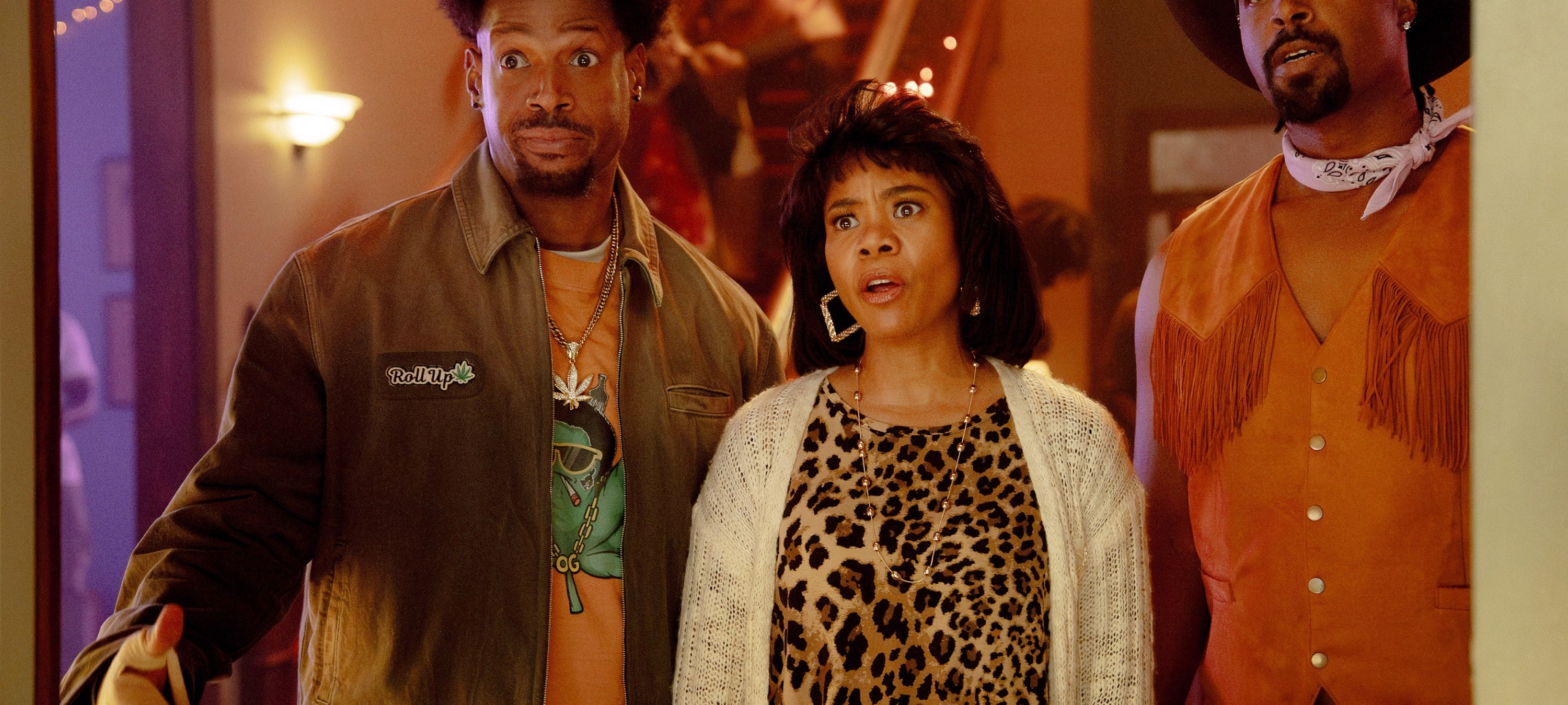 Three people stand indoors at a party, looking surprised. One wears a jacket, another a leopard print outfit, and the third a cowboy-style vest and hat