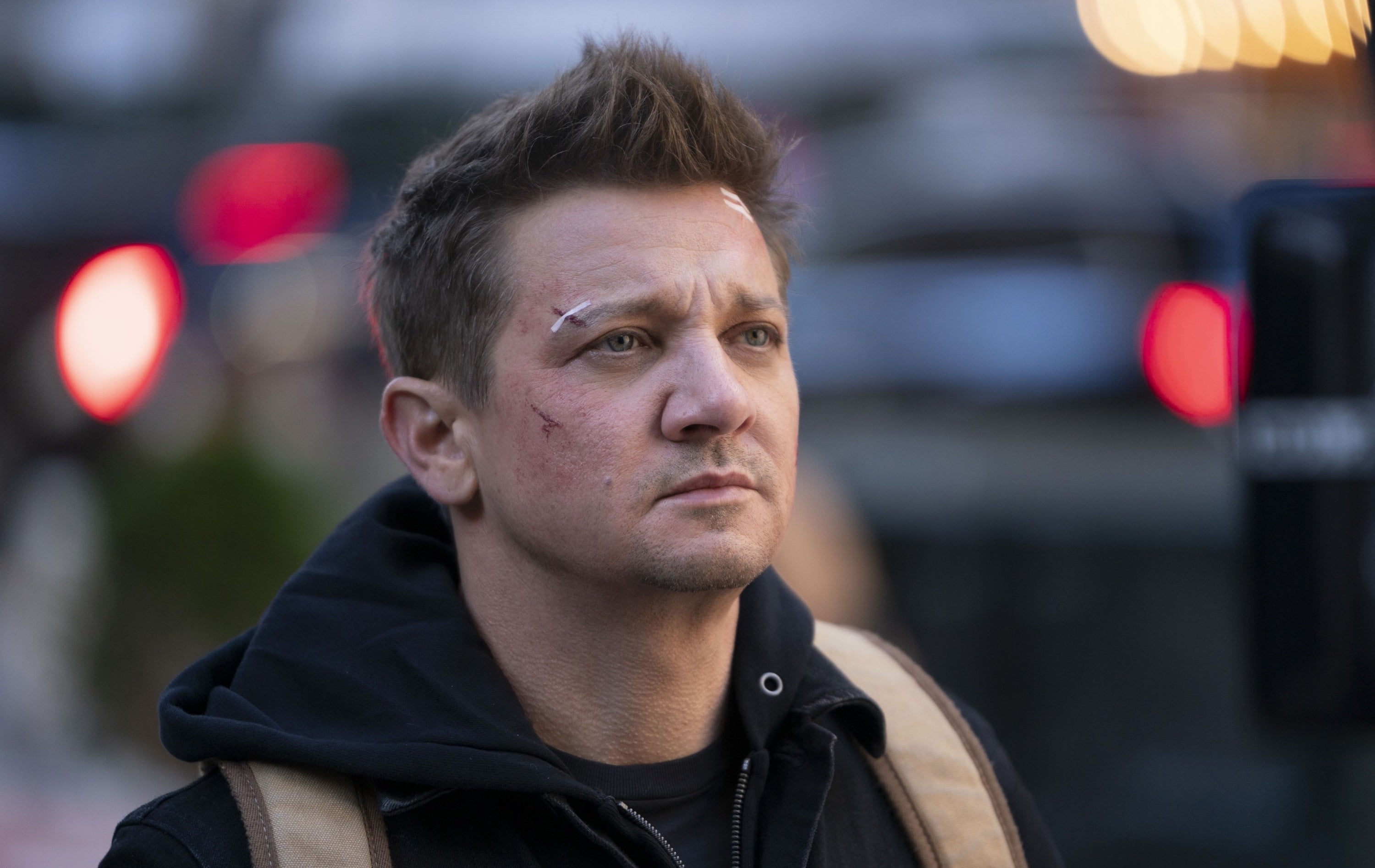 A person with short hair and facial cuts wears a hoodie and backpack, looking into the distance