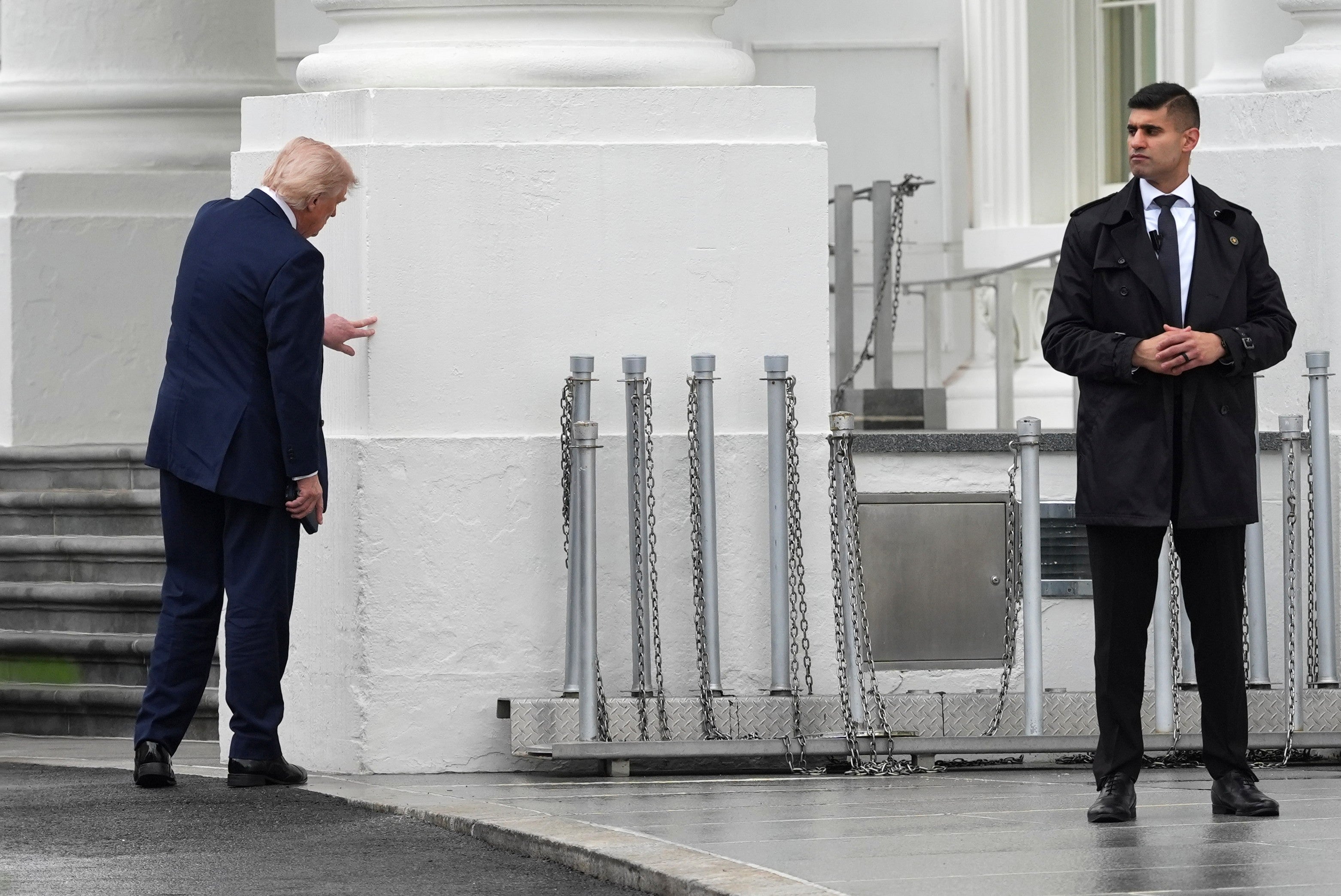 En person i dress gestikulerer mot en vegg utenfor en stor bygning, mens en annen person i sikkerhetsuniform står i nærheten