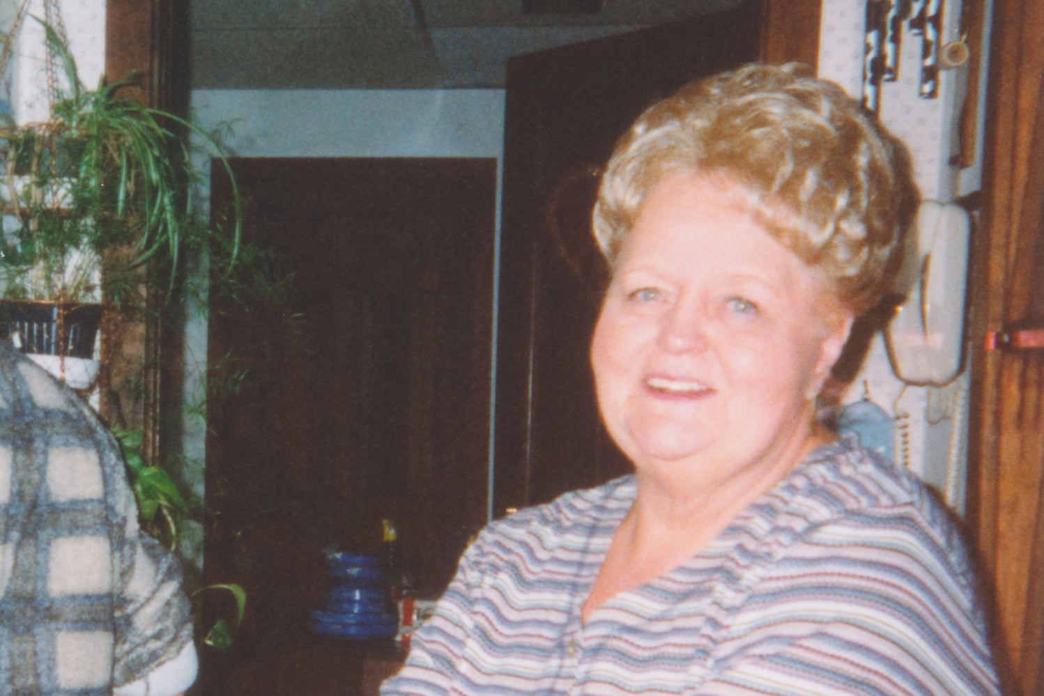 An older woman with a beehive hairstyle stands in a cozy room, smiling with arms crossed, wearing a striped top