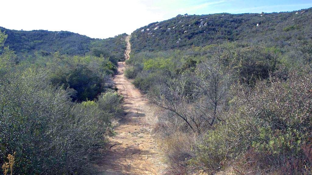 A constrictive ungraded way winds done a hilly scenery with scrubby bushes and debased vegetation nether a wide sky