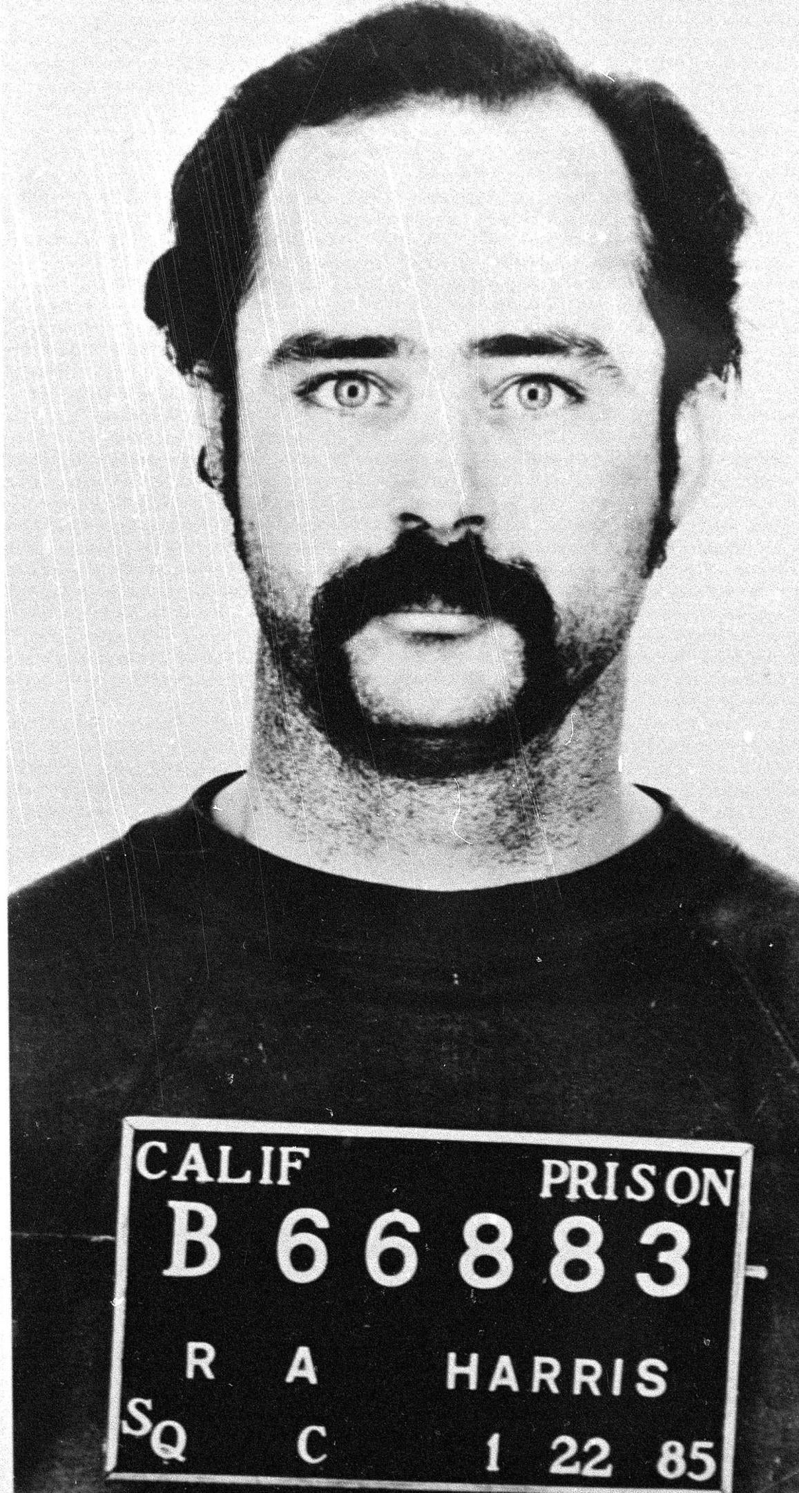 A achromatic and achromatic mugshot of a antheral with a mustache, wearing a situation azygous labeled "Calif Prison B66883" dated 1985