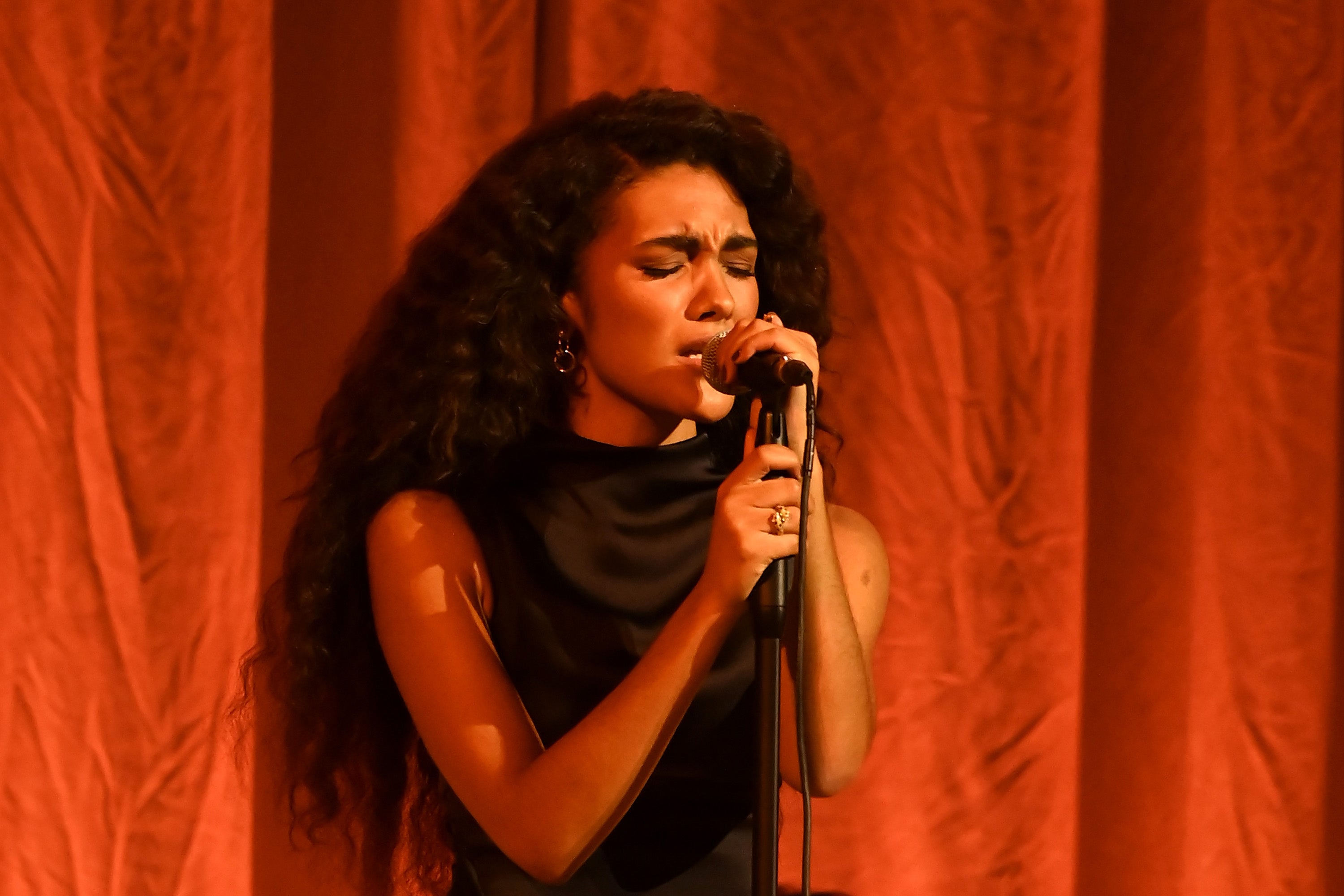 A person with long hair performs on stage, passionately singing into a microphone, wearing an elegant black outfit