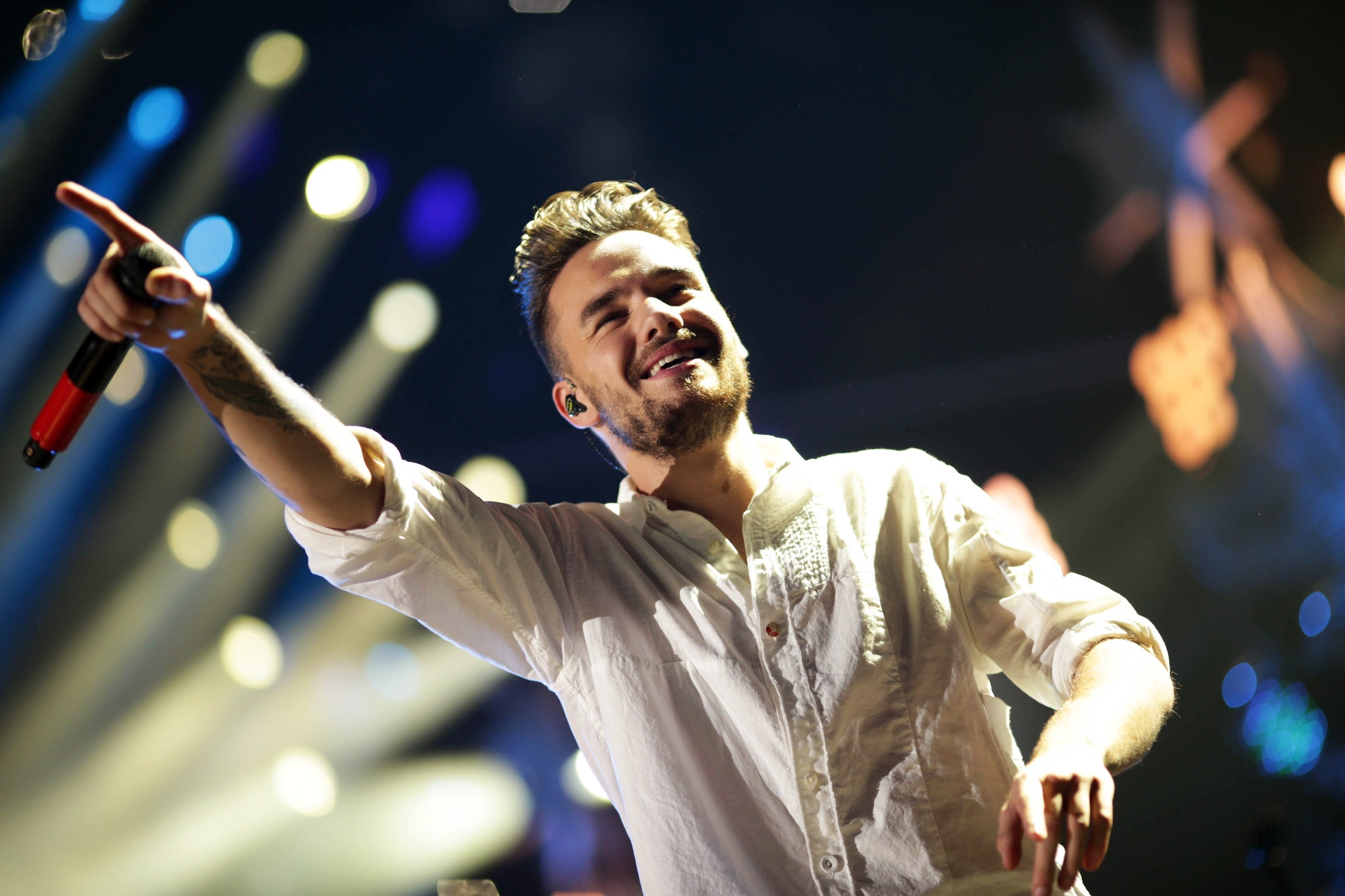 Singer performing energetically on stage, wearing a casual white shirt, holding a microphone, and pointing forward. Stage lights illuminate the background