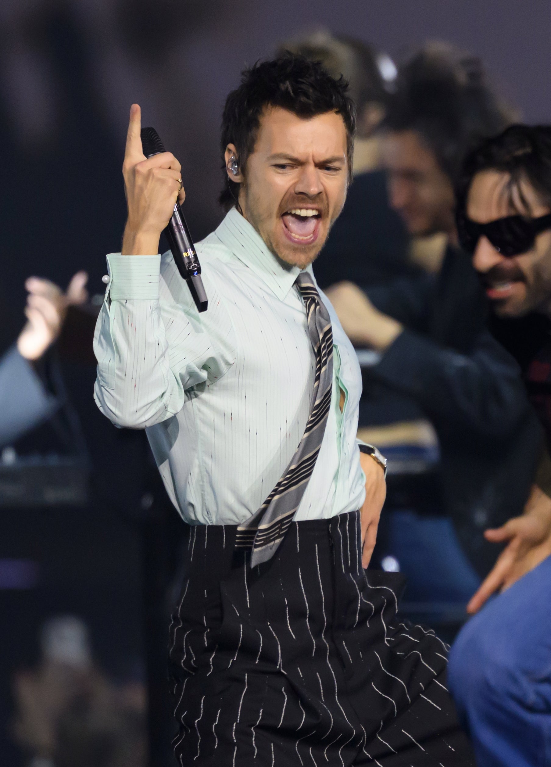 A singer energetically performs on stage, wearing a pinstripe suit with a striped shirt and tie