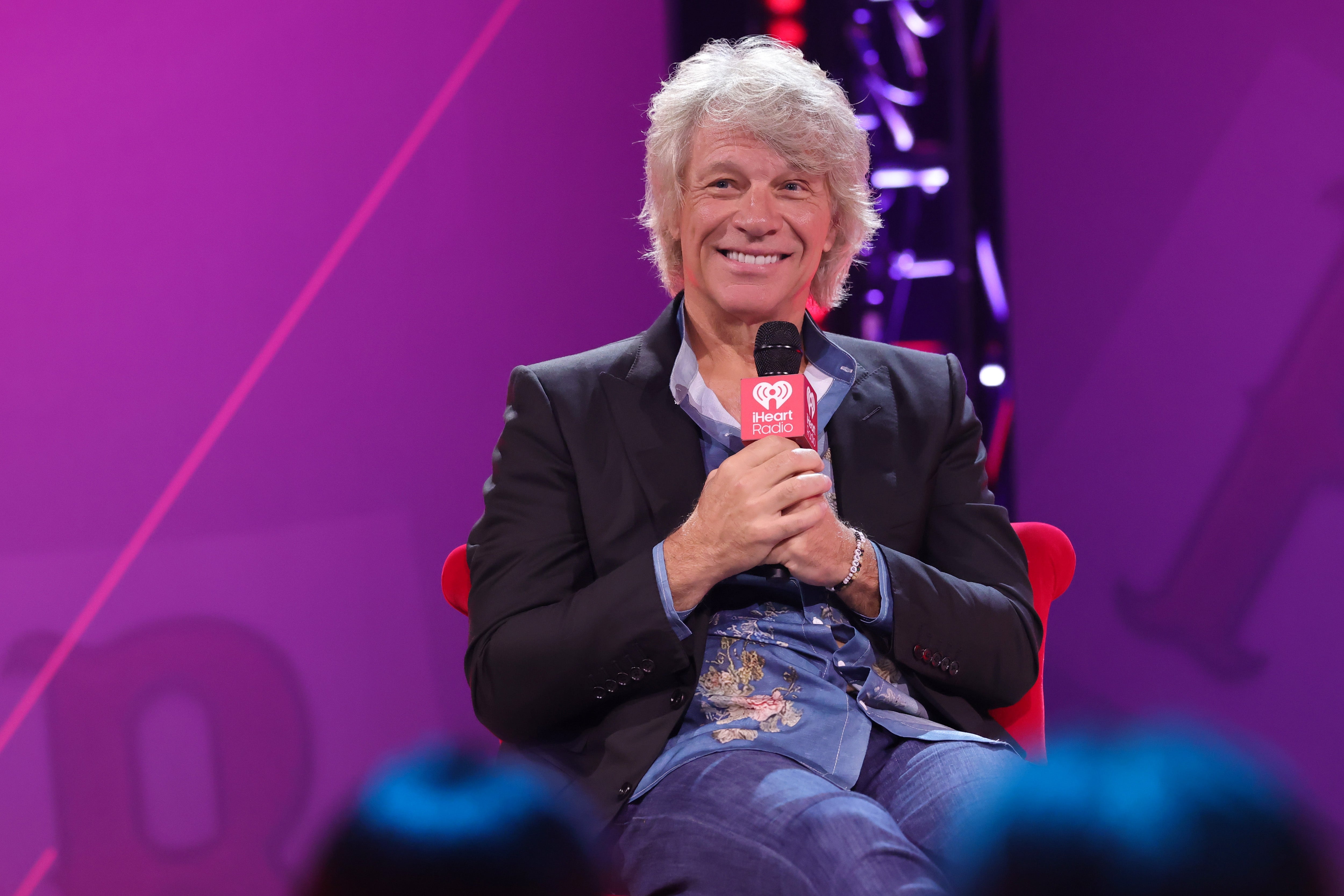 Celebrity sitting on stage, smiling while holding a microphone. Dressed casually with a patterned shirt