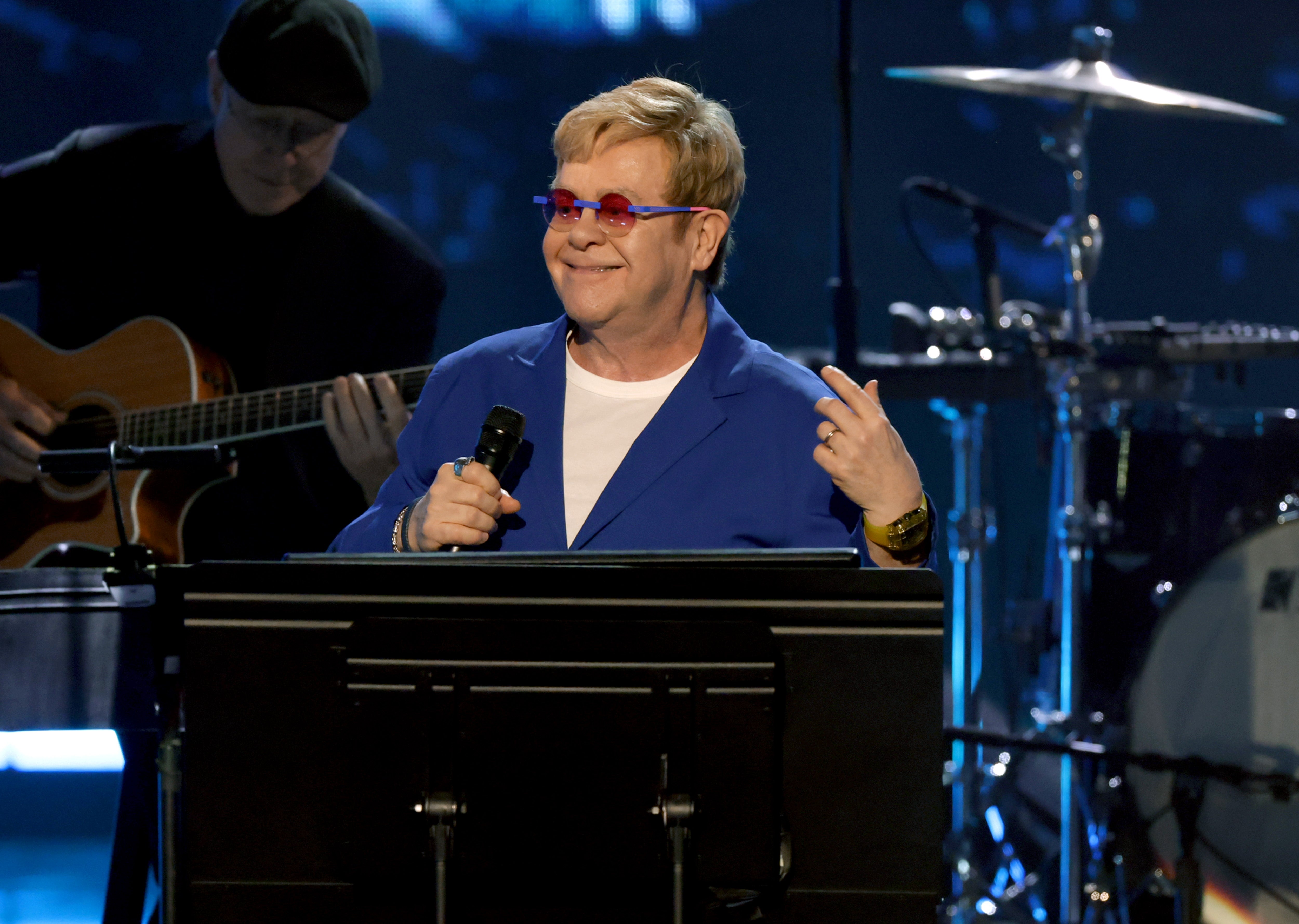 A person on stage wearing a blue suit and red glasses, smiling while performing, with musicians in the background