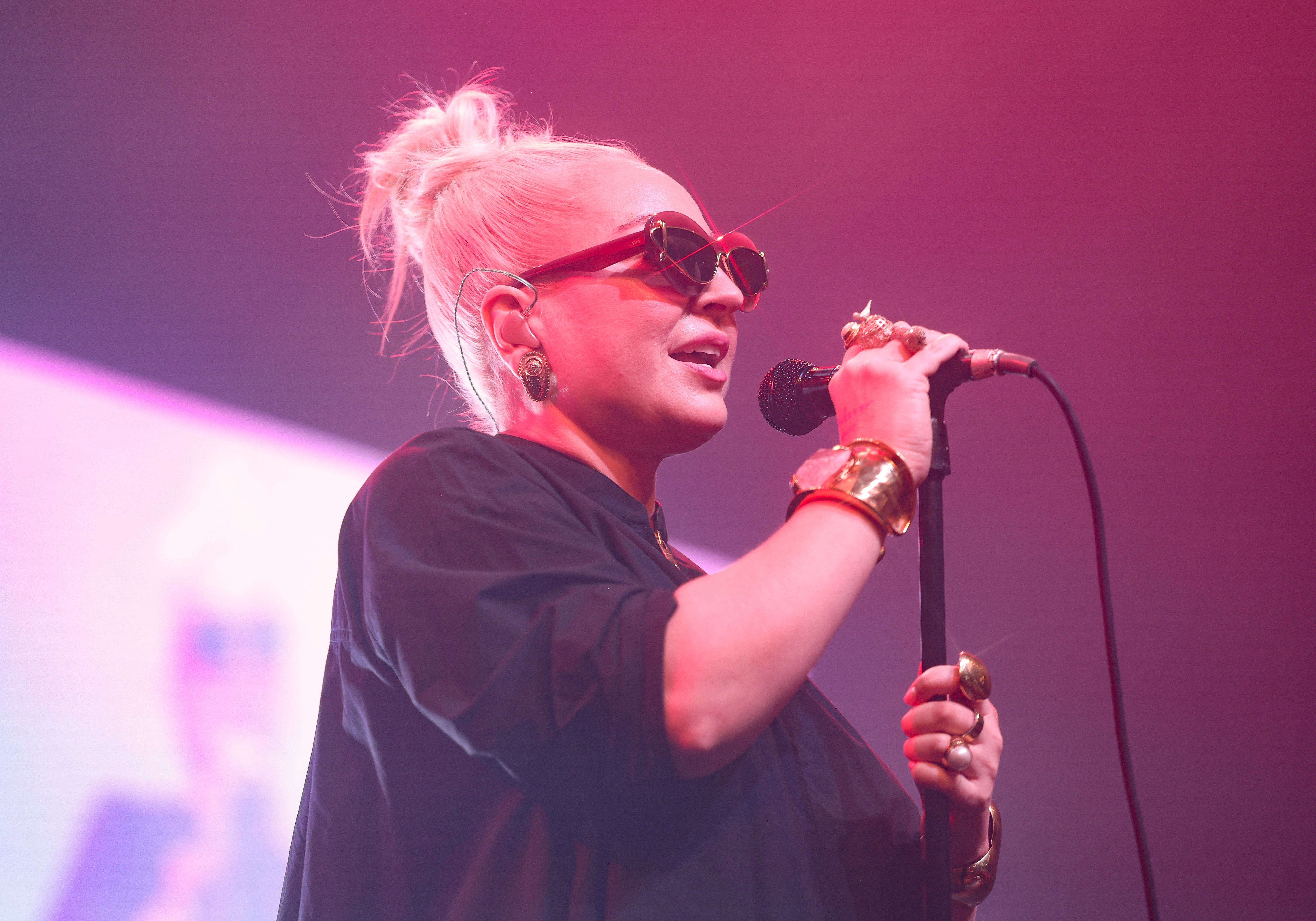 A person performs on stage wearing oversized sunglasses, a black top, and chunky gold jewelry, holding a microphone against a blurred background