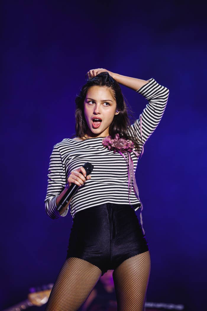A performer sings energetically on stage, wearing a striped top and high-waisted shorts, holding a microphone