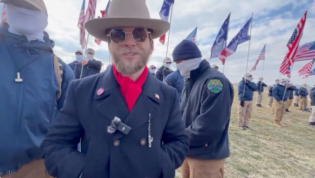 A antheral   successful  a wide-brimmed chapeau  and sunglasses is surrounded by masked individuals holding assorted  flags outdoors