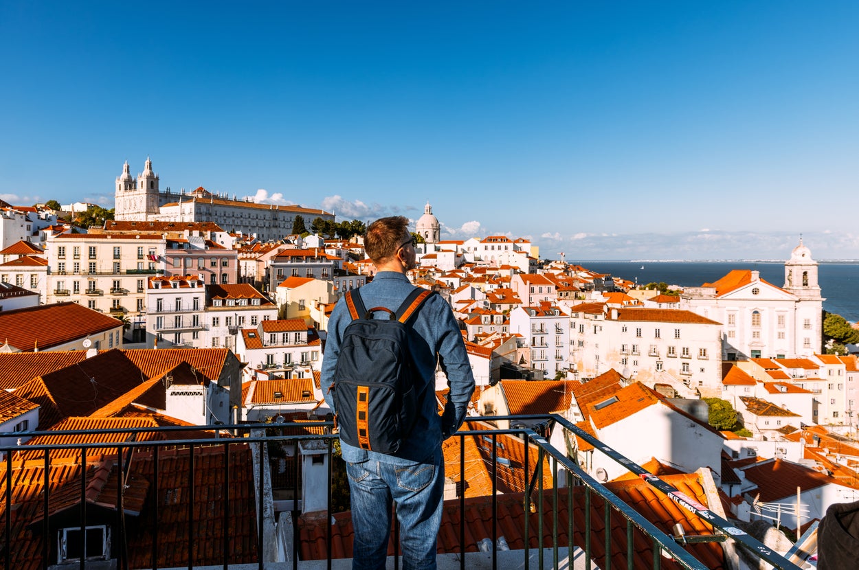 Person with a backpack stands on a balcony overlooking a scenic cityscape with historic buildings and a distant sea view