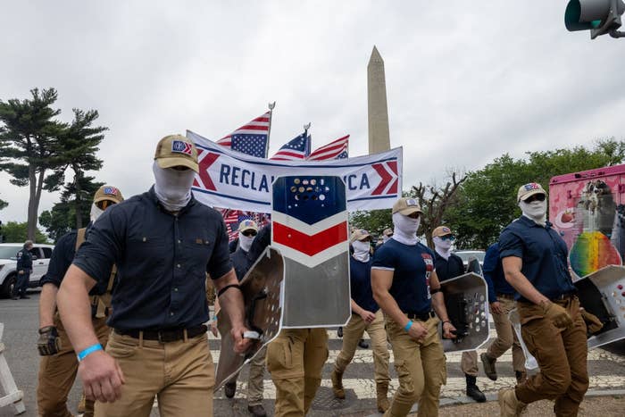 Group of masked individuals successful  matching attire march with shields adjacent   the Washington Monument, holding "Reclaim America" banner
