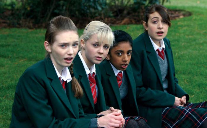 Four students in school uniforms, sitting on grass, look towards the camera with expressions of surprise or concern