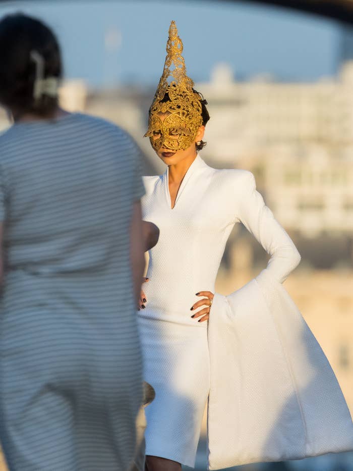 Person in an elegant white outfit with dramatic sleeves and a detailed golden face mask, standing outdoor