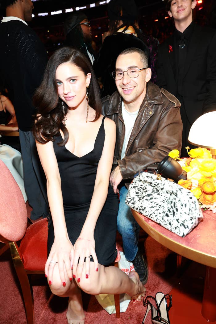 Two radical   seated; the pistillate   wears a chic achromatic  dress, the antheral   sports a casual leather jacket. They airs  astatine  a glam lawsuit   with others successful  the background