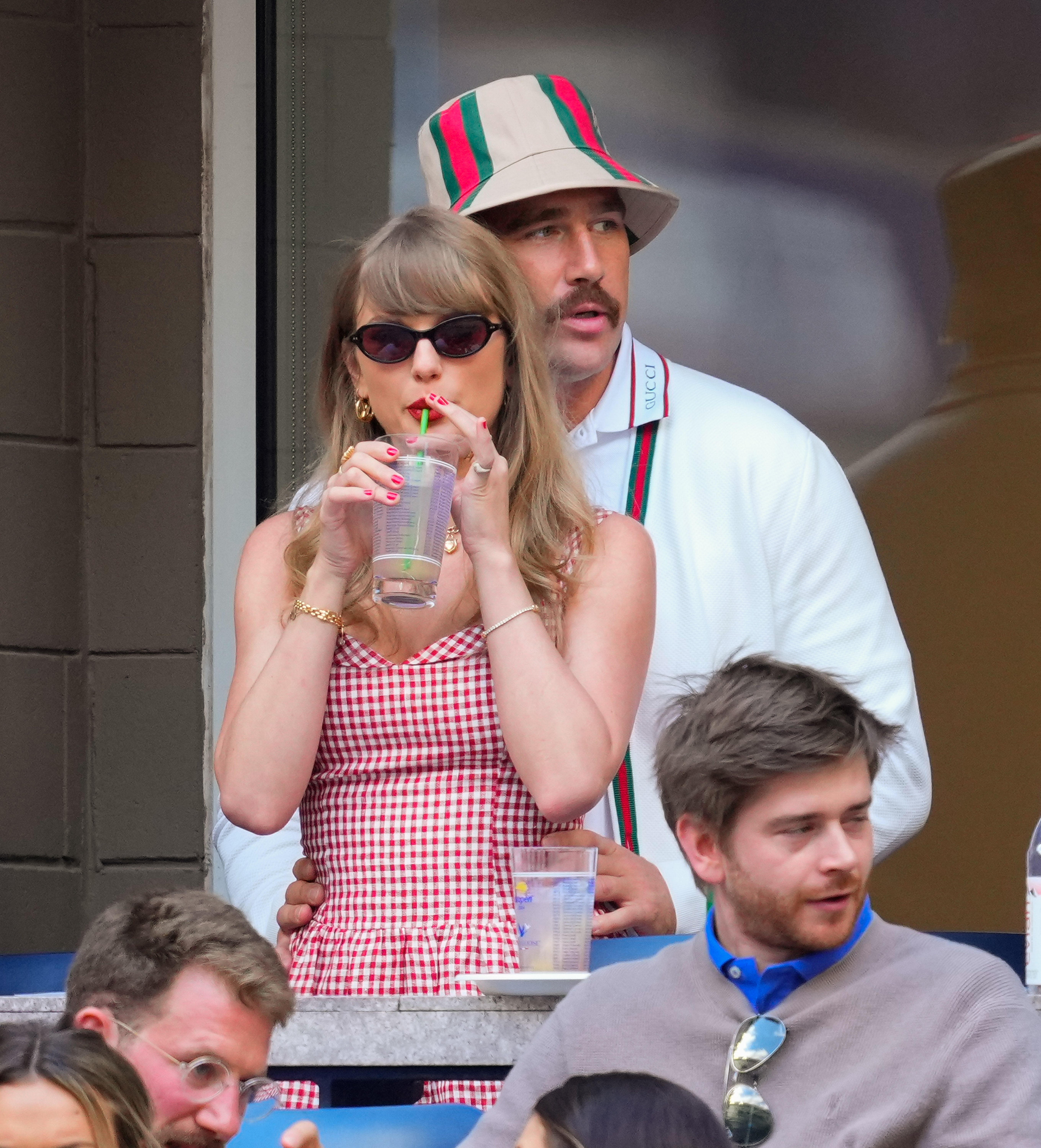 Taylor Swift successful a gingham formal and sunglasses sips a drink, with Travis Kelce successful a bucket chapeau and sporty attire lasting down her astatine an event