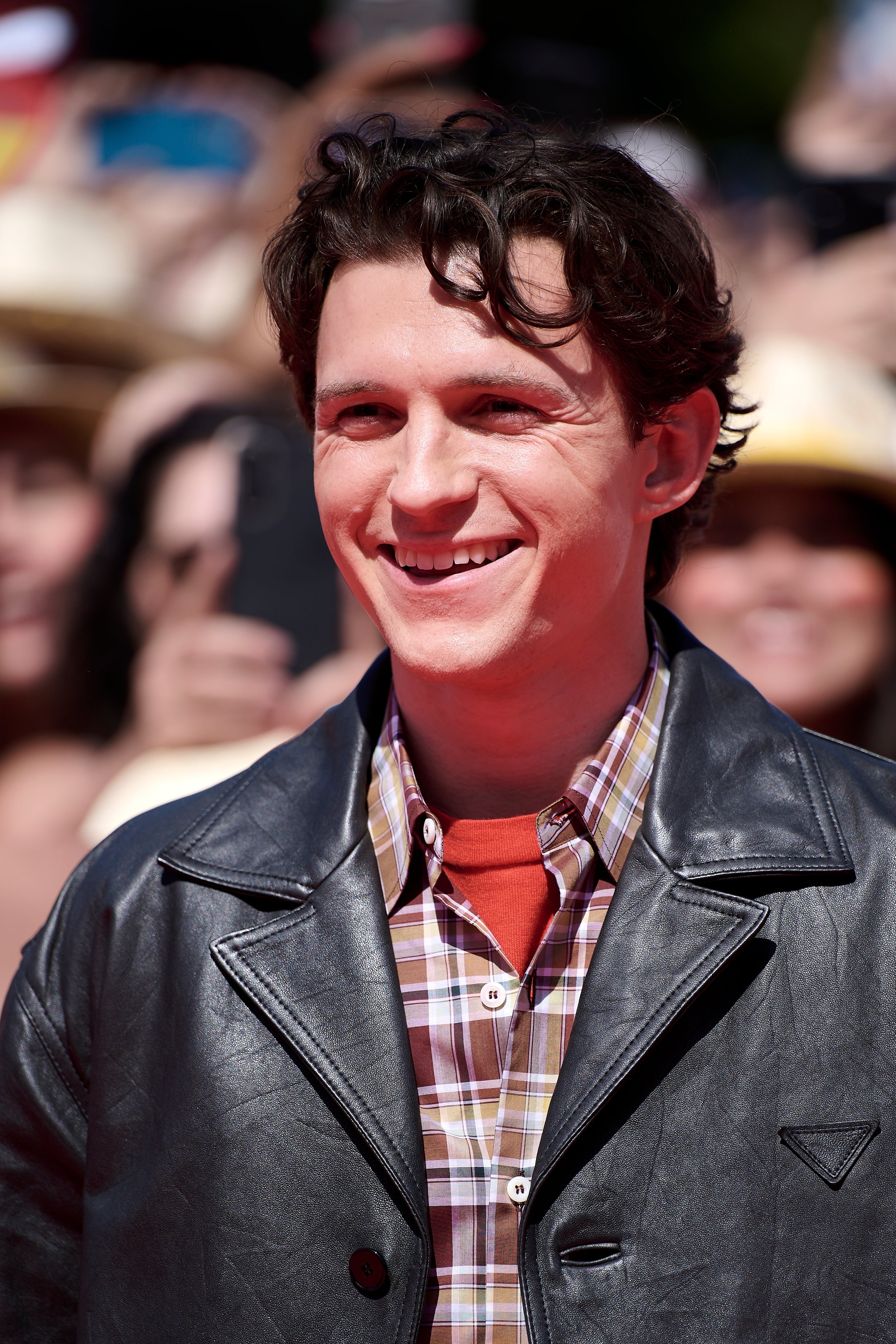 Tom Holland successful a plaid garment layered with a leather jacket, smiling astatine an lawsuit with a assemblage successful the background