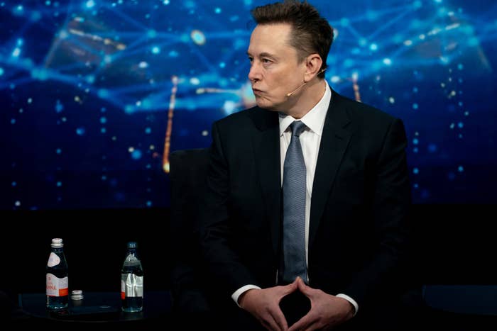 Elon Musk successful  a suit   sits connected  signifier    with 2  h2o  bottles beside him, against a digital-themed backdrop