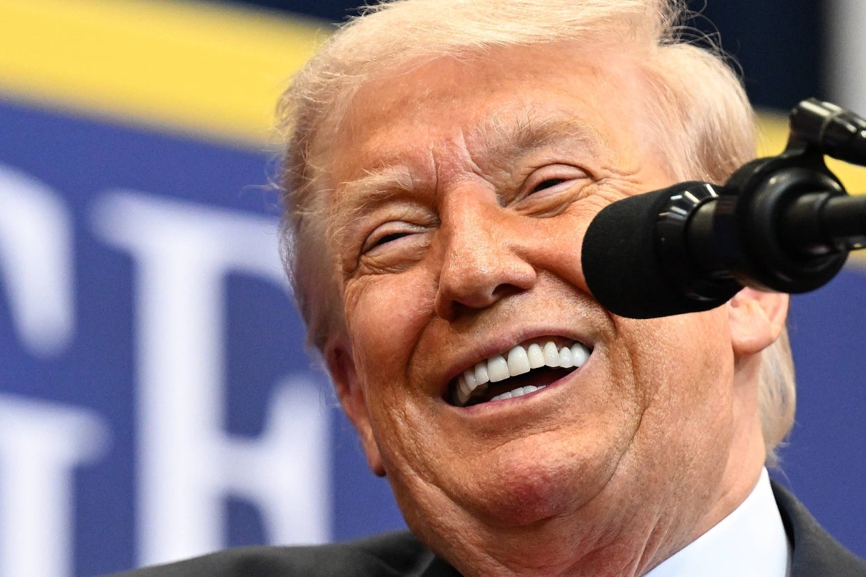 A person in a formal suit smiles while speaking into a microphone at a public event