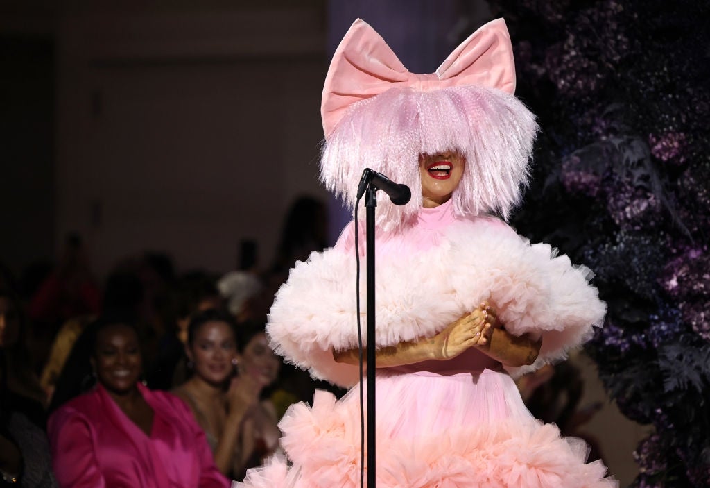 Celebrity in a voluminous pink dress and large bow headpiece performs on stage, with audience visible in the background