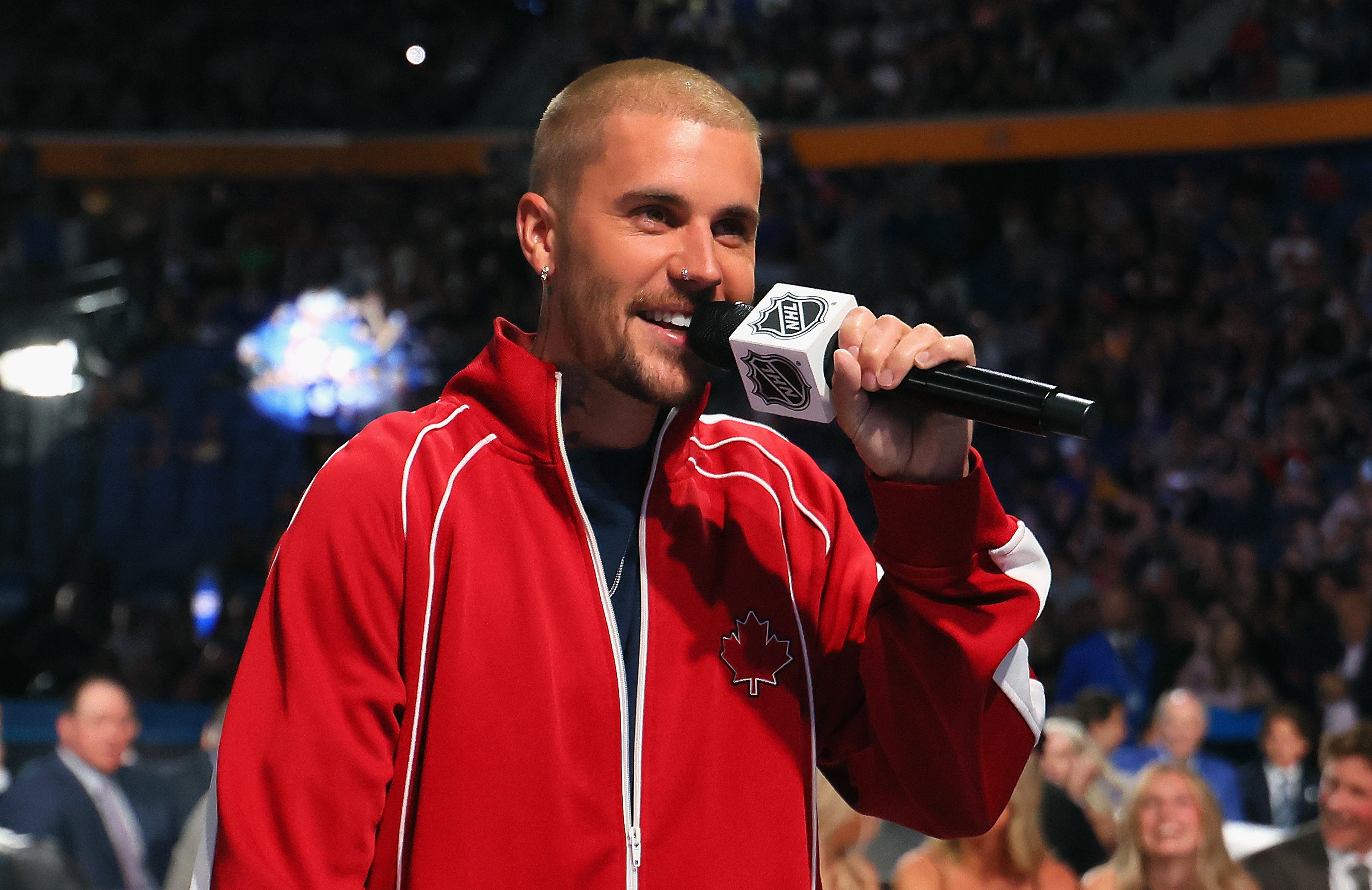 Man in a casual red track jacket with a maple leaf emblem speaks into a microphone at an event