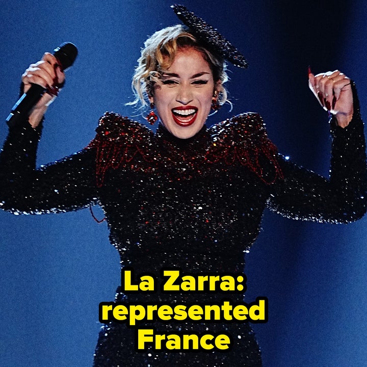 Singer on stage in a sparkling gown performs energetically, holding a microphone with arms raised, against a backdrop of spotlight beams